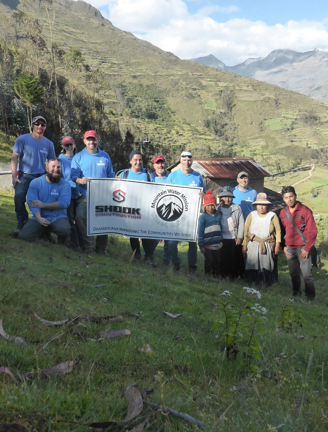 A group of people pose outdoors on a grassy hillside holding a banner. Mountains and a rustic building are in the background, with blue sky and clouds above.
