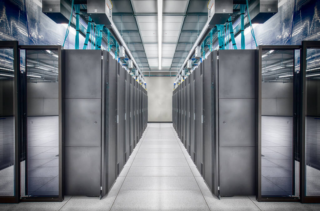 A modern data center with rows of black server racks on both sides of a bright, clean aisle, under a high ceiling with overhead cabling and fluorescent lights.
