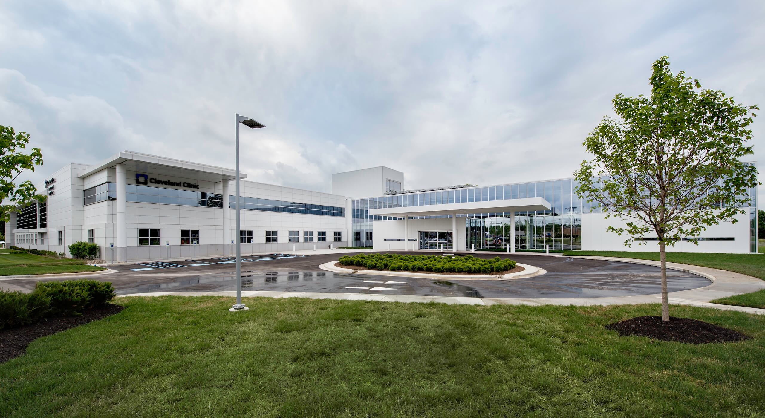A modern white medical building with large windows, labeled Cleveland Clinic, sits under a cloudy sky. The scene includes a driveway, parking spaces, green grass, and a few trees.