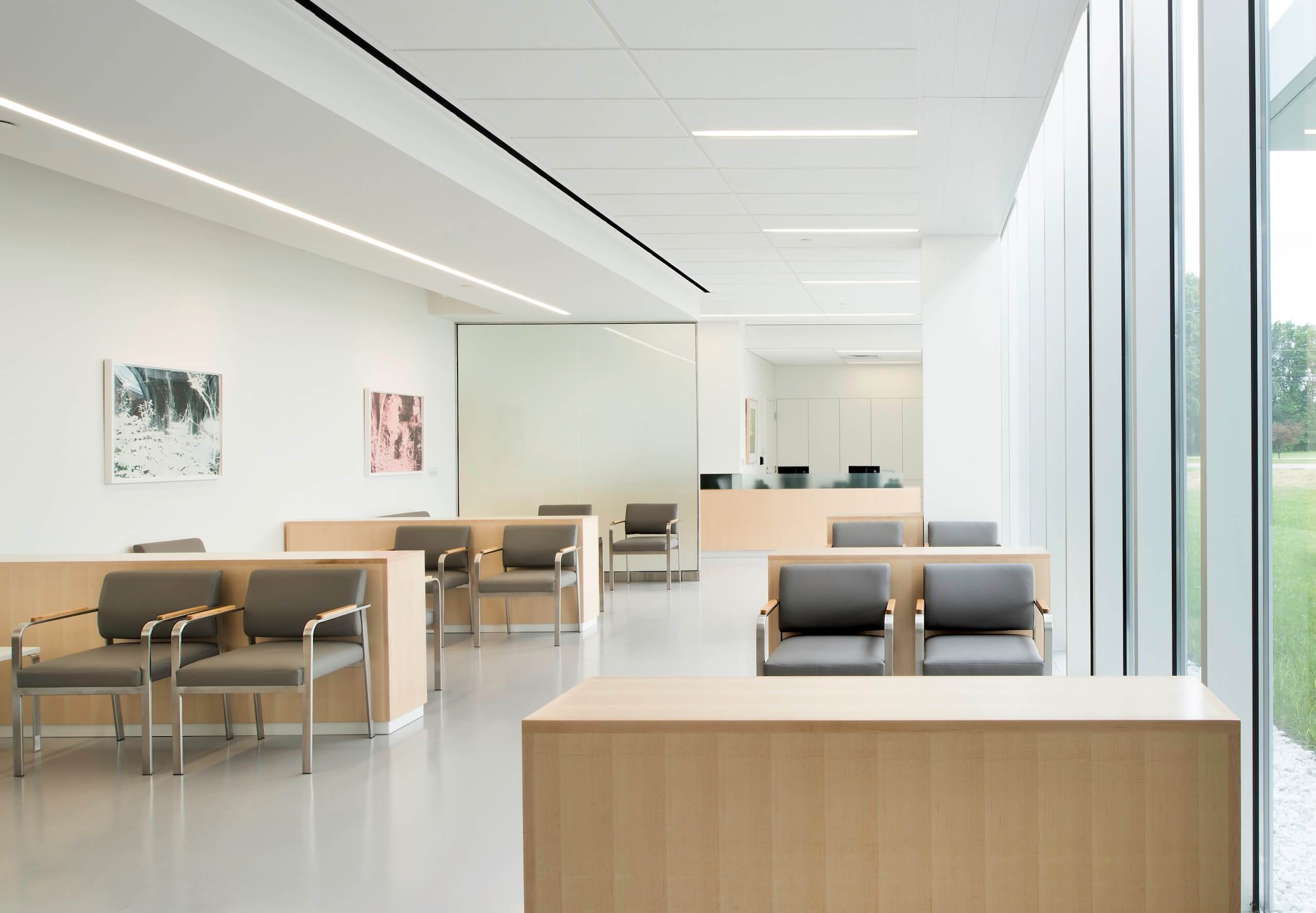 A modern, bright waiting room with gray chairs, light wood accents, large windows, white walls, and minimal artwork. The space appears clean, orderly, and quiet, with a view of greenery outside.