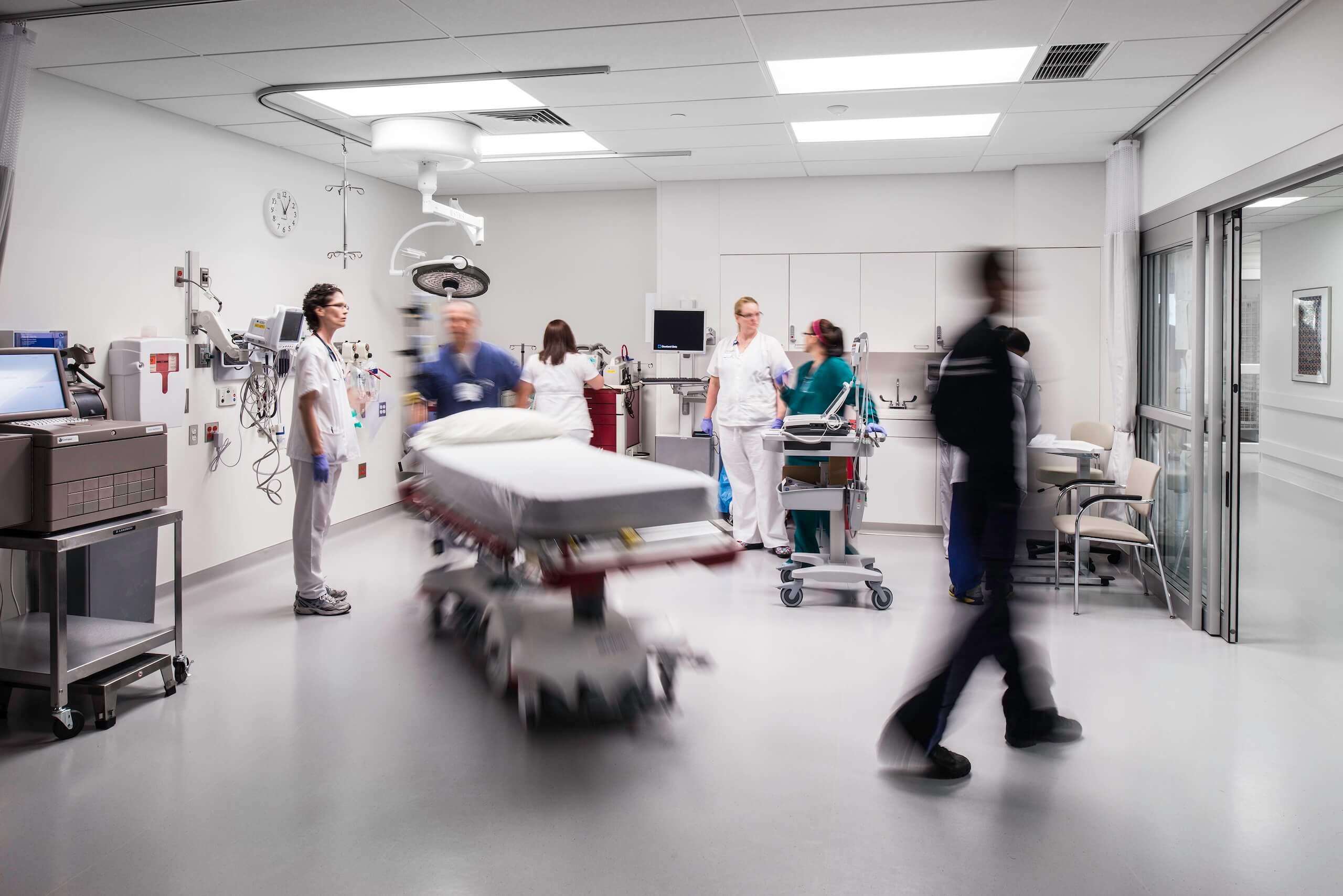 Doctors and nurses stand around a hospital bed in a bright emergency room as a blurred figure walks by, conveying urgency and activity.