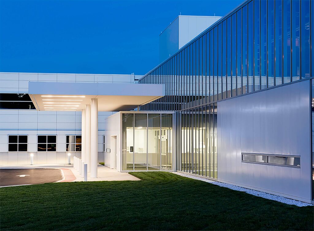 A modern building with large glass windows and a well-lit entrance, photographed at dusk. The exterior features sleek lines and a manicured lawn in front.