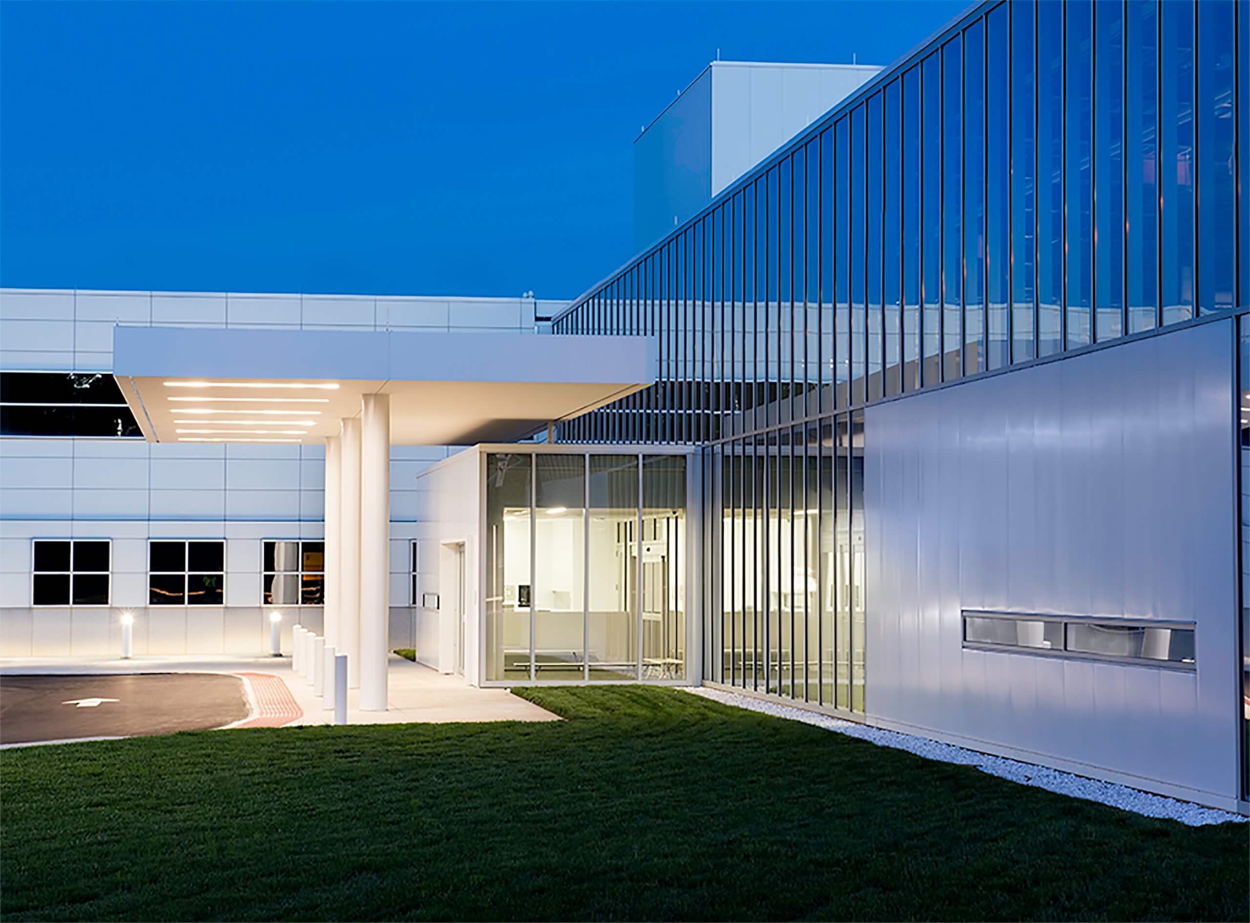A modern building with large glass windows and a well-lit entrance, photographed at dusk. The exterior features sleek lines and a manicured lawn in front.