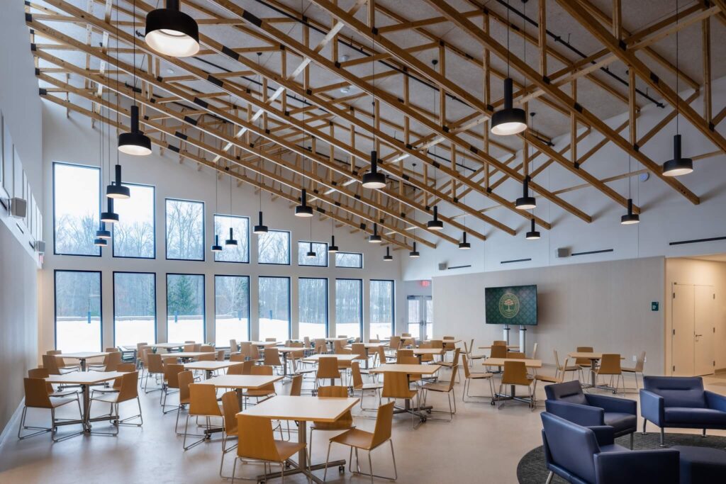Spacious cafeteria with wooden tables and chairs, large windows letting in natural light, exposed wooden ceiling beams, modern light fixtures, and a TV mounted on the wall. Snowy landscape visible outside.
