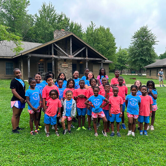 A group of children and a few adults pose together on green grass in front of rustic cabins, wearing matching blue and pink camp shirts, outdoors on a cloudy day.