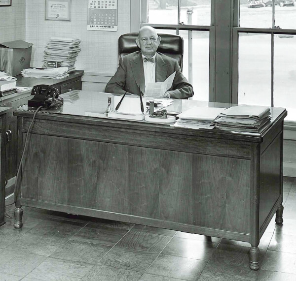 An older man in a suit sits at a large wooden desk in an office, holding papers. The desk is covered with documents, a rotary phone, and office supplies. Large windows and stacked papers are in the background.