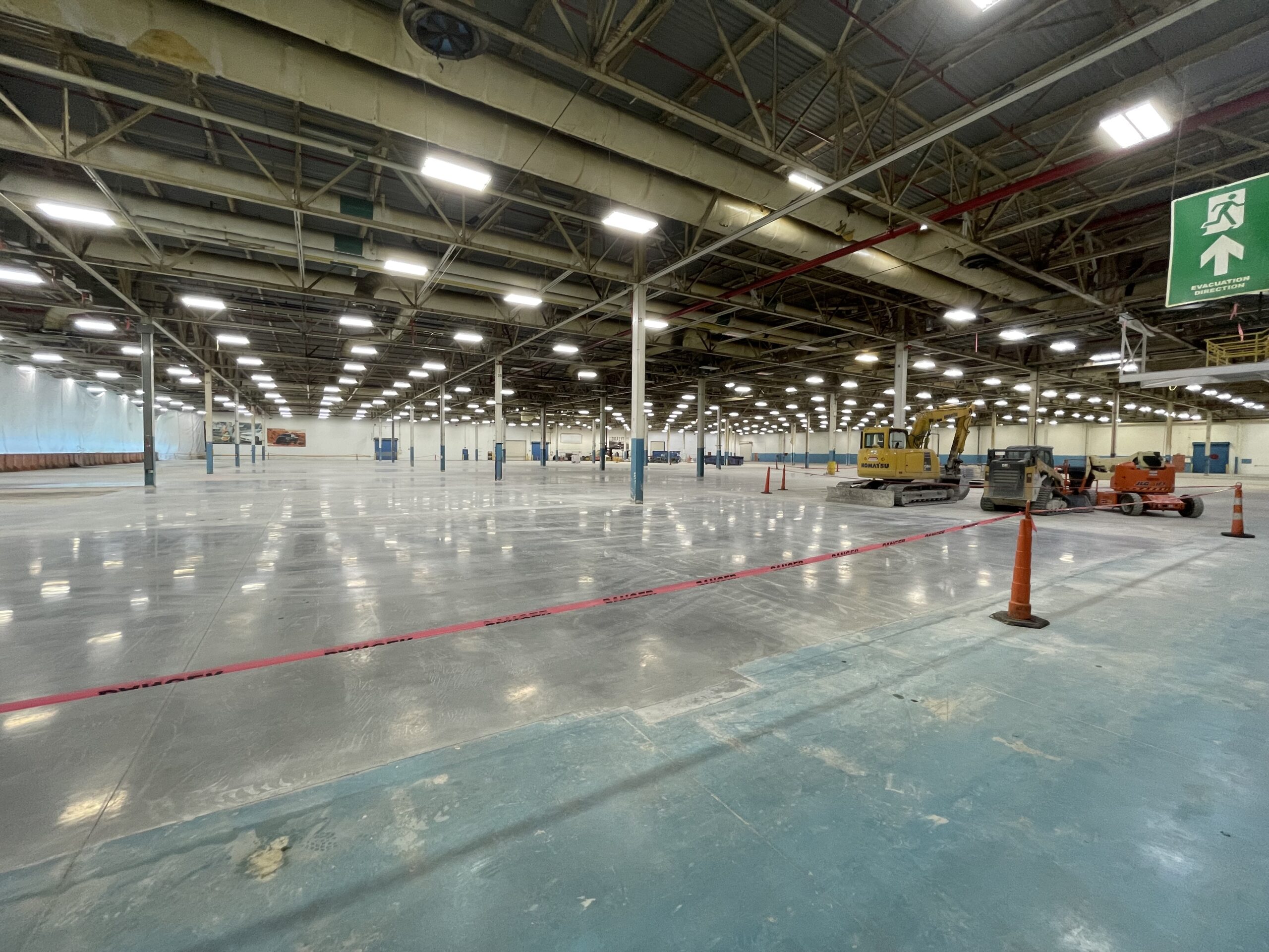 A large, well-lit industrial warehouse with a polished concrete floor, construction equipment, orange cones, and red caution tape. Overhead lights and structural beams are visible.