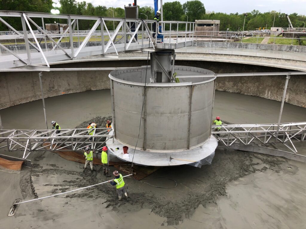 Construction workers in safety vests and helmets install a large metal structure in a circular concrete basin, using scaffolding and tools, at an outdoor industrial site with trees in the background.