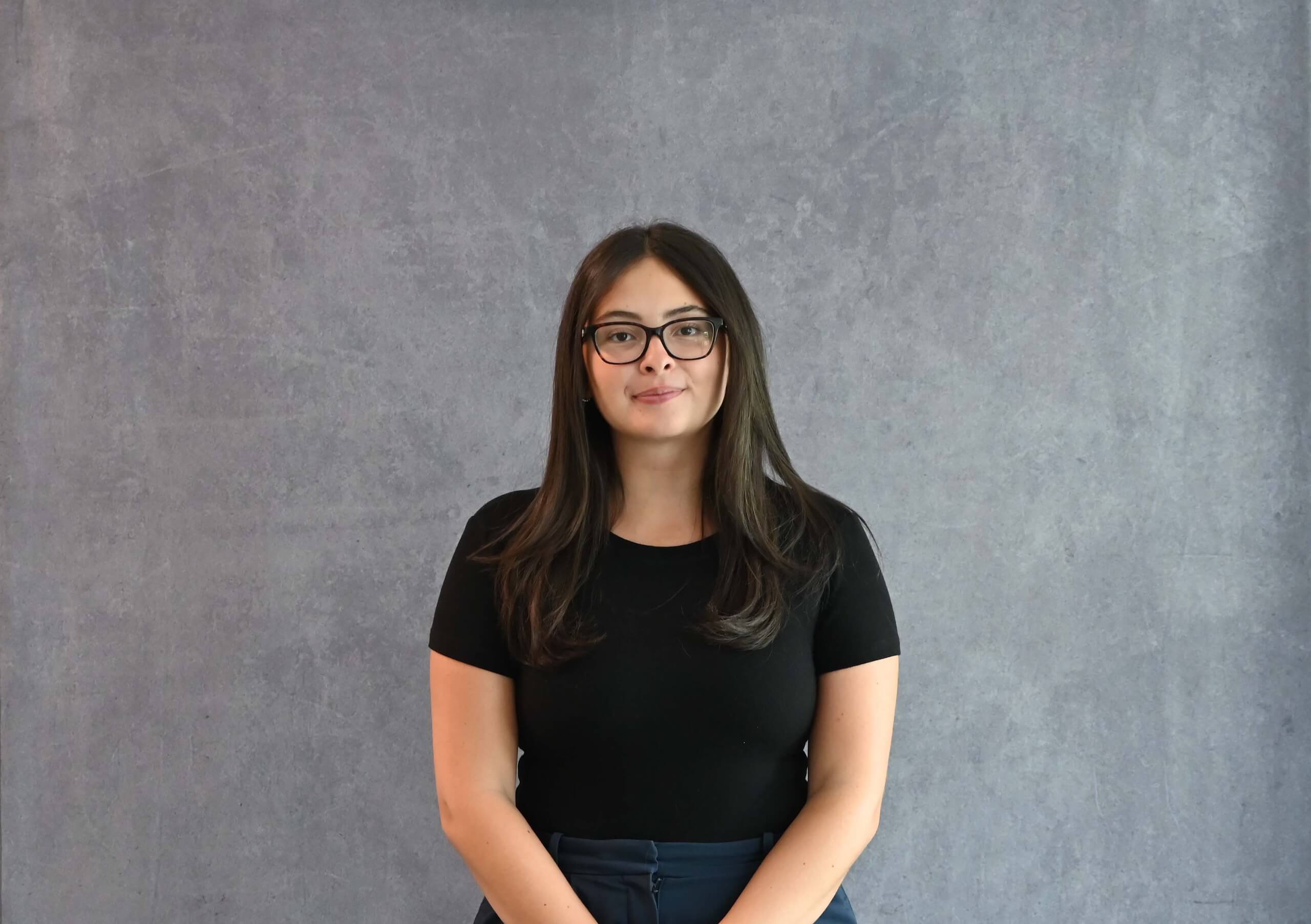 A woman with long brown hair and glasses, wearing a black shirt, stands in front of a gray textured background, facing the camera with a neutral expression.