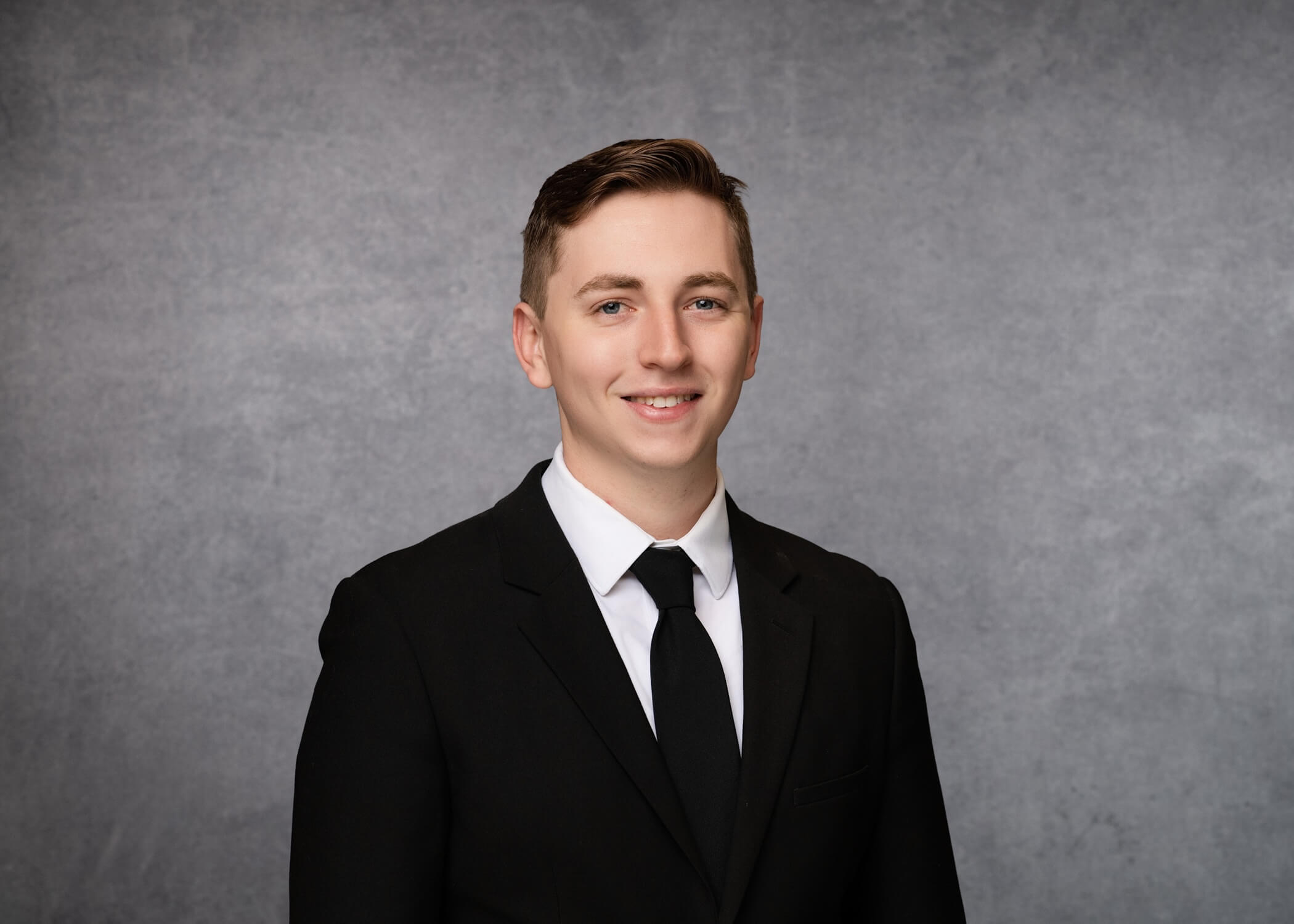 Young man in a black suit, white shirt, and black tie, smiling in front of a plain gray backdrop.