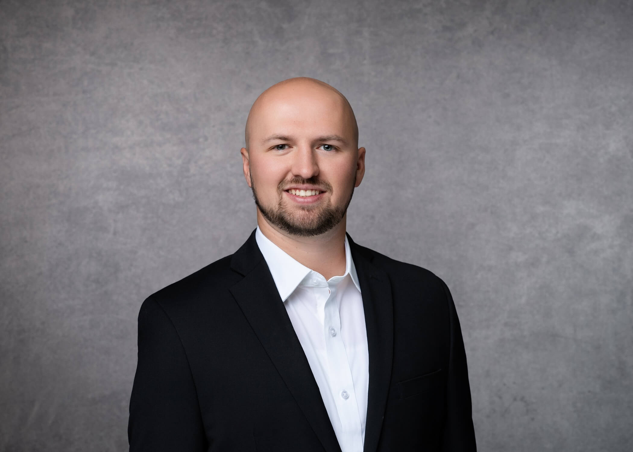 A man in a black suit jacket and white shirt stands smiling in front of a plain gray background.