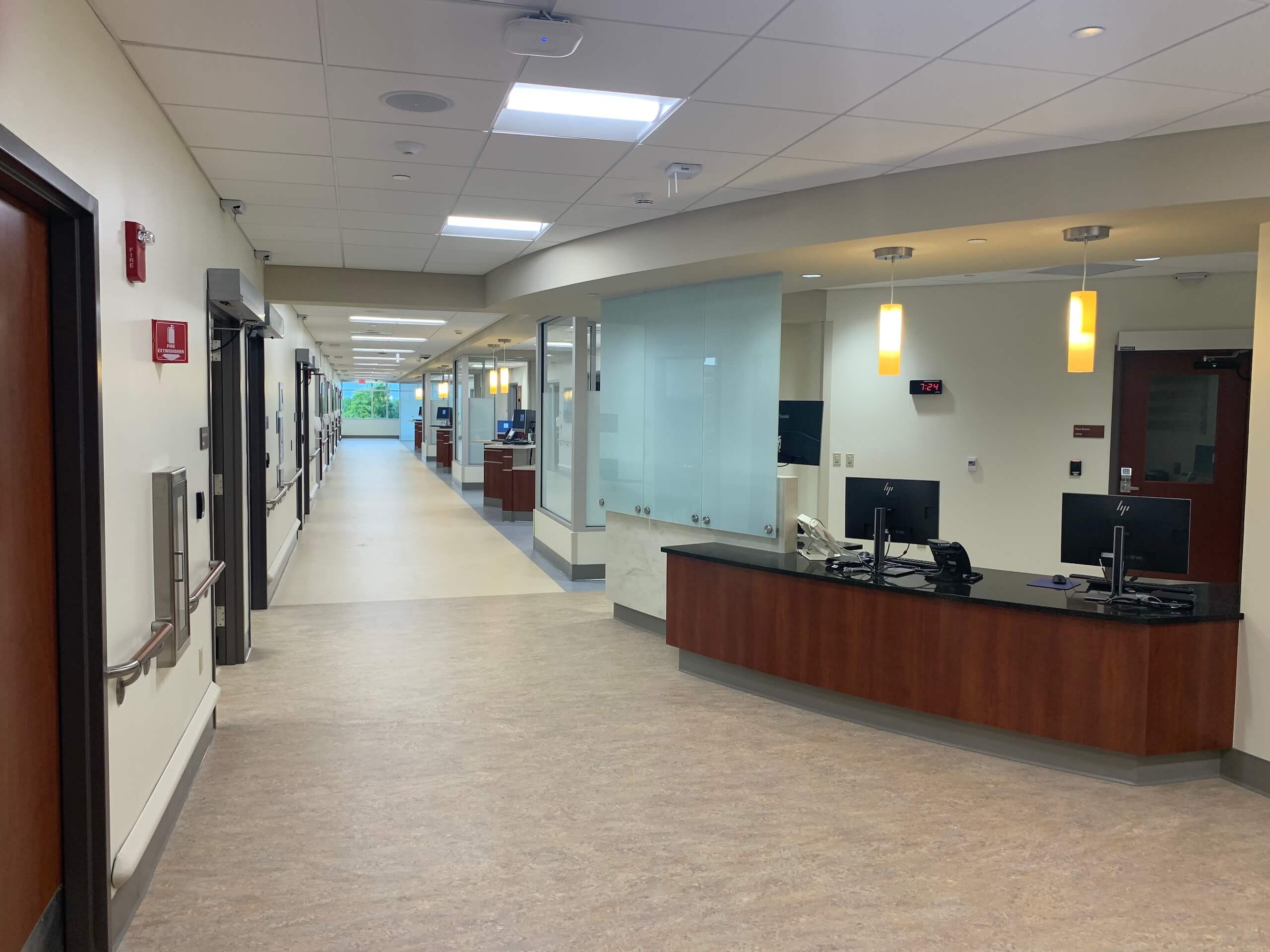 A clean, modern hospital hallway with a nurses station on the right. Two computer monitors sit on the desk, and patient rooms line the hallway. Overhead lights brightly illuminate the area.