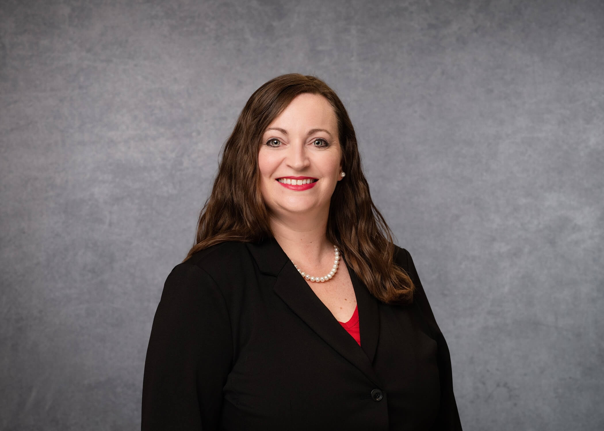 A woman with long brown hair, wearing a black blazer, red top, and pearl necklace, smiles in front of a plain gray background.