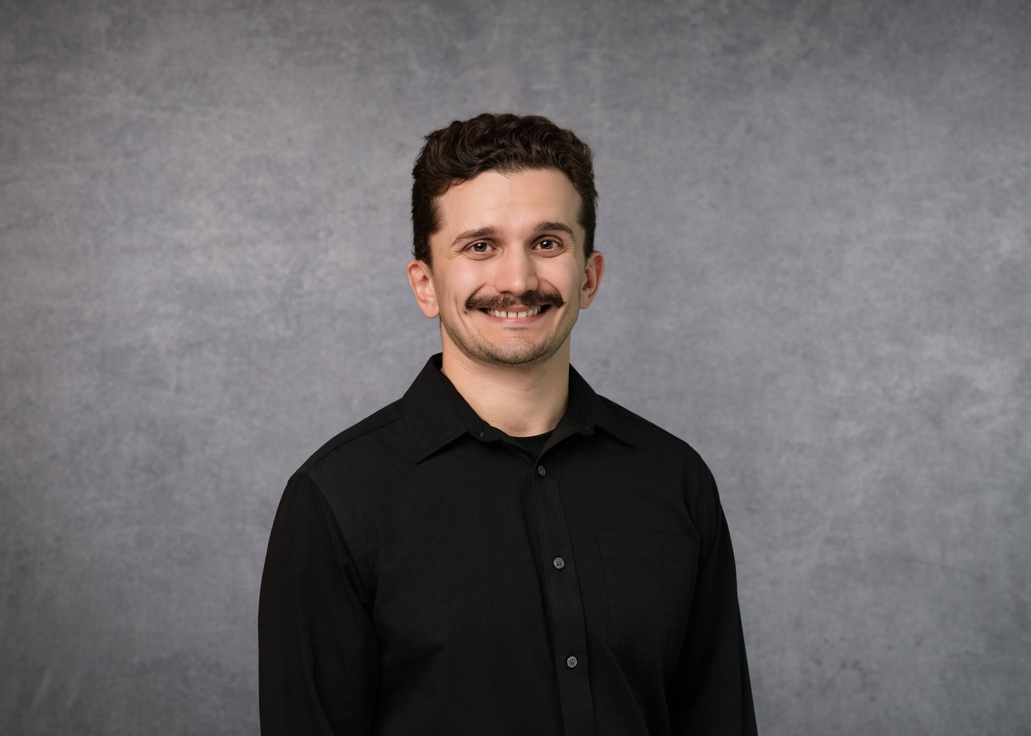 A man with short curly hair, a mustache, and a black button-up shirt smiles at the camera against a plain gray background.