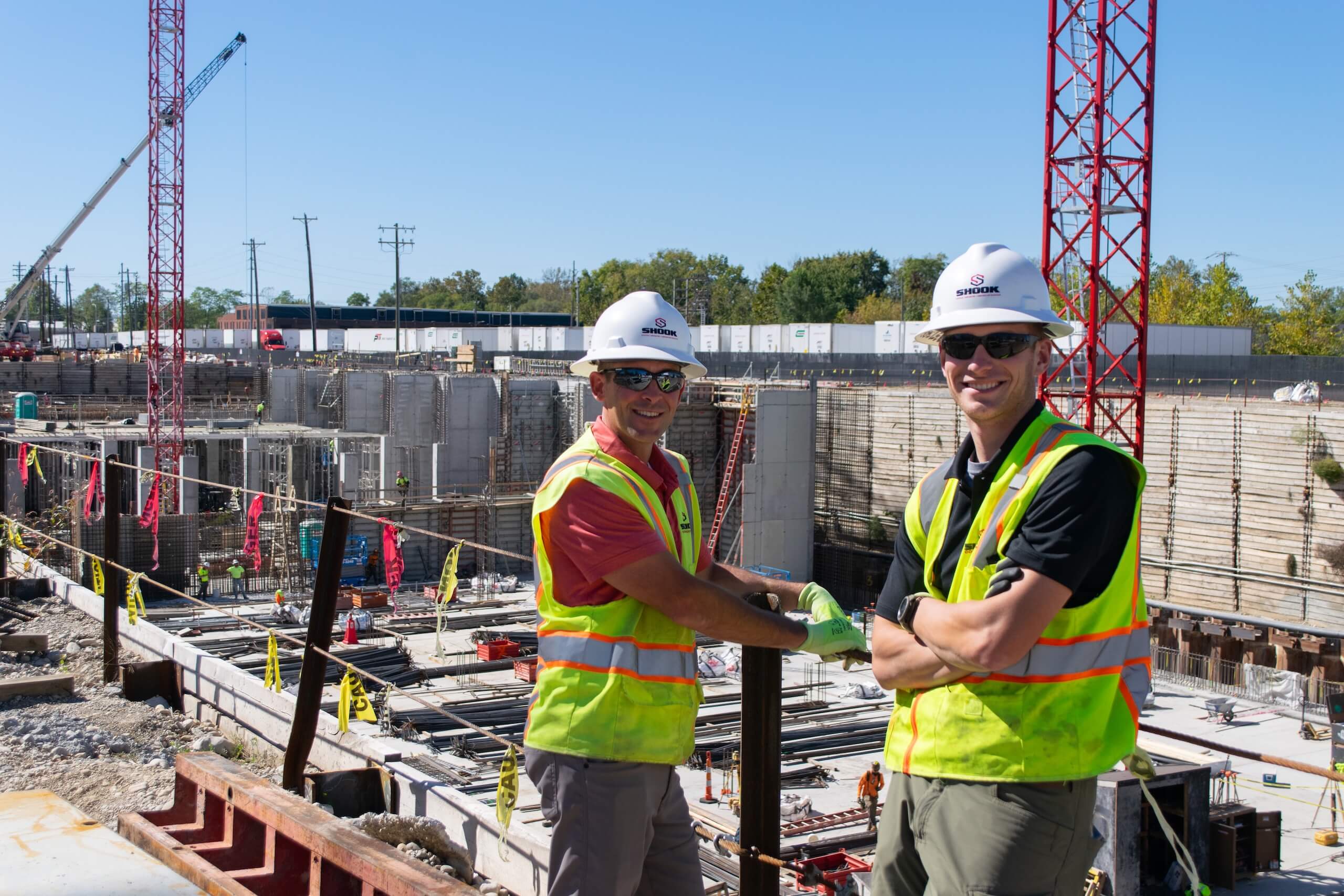 Two construction workers in safety vests and hard hats smile at the camera while standing at an active construction site with cranes and building materials in the background.