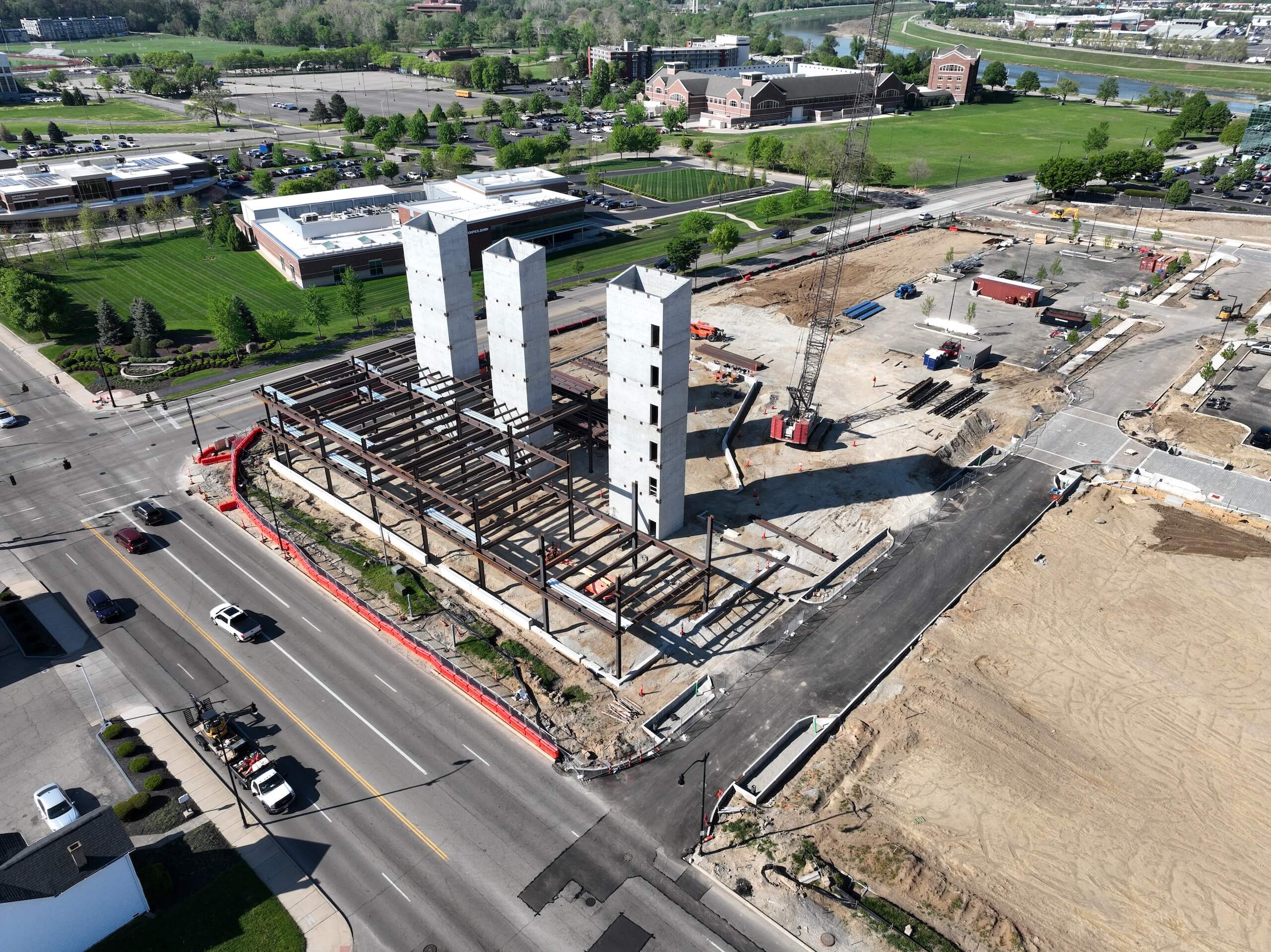 Aerial view of a building under construction with steel framework and three tall concrete elevator shafts. Nearby roads, green lawns, and other buildings are visible. Construction vehicles and equipment are present on site.