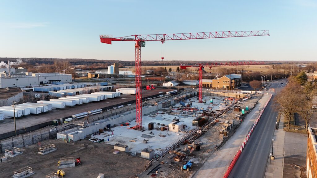 Aerial view of a construction site with two large red cranes, building foundations, construction materials, nearby industrial buildings, and a road running alongside the site. Trees and an open landscape are in the background.