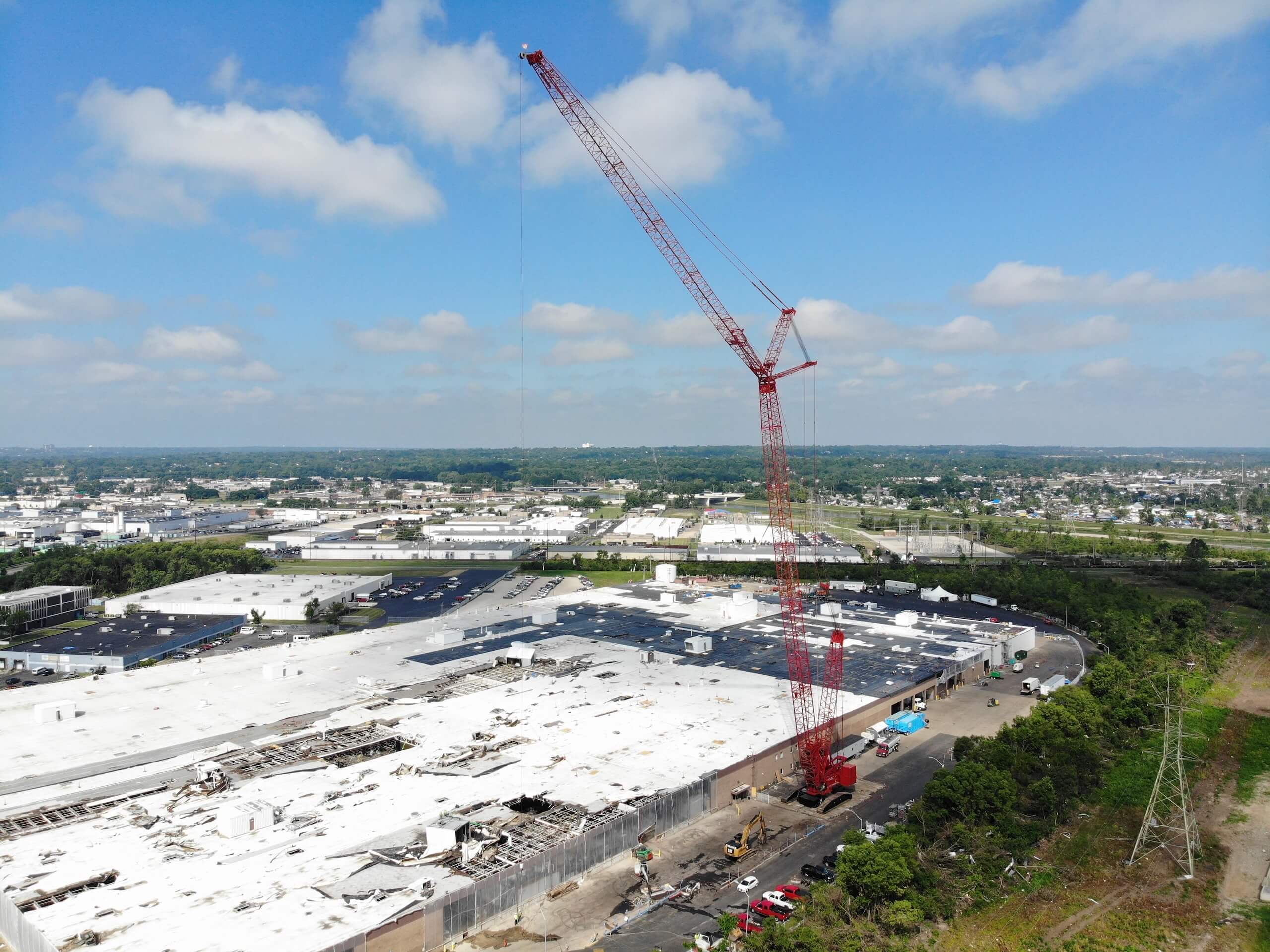 A large red crane operates beside a white industrial building under construction or demolition, with vehicles and equipment nearby, set against a suburban landscape and blue sky with scattered clouds.