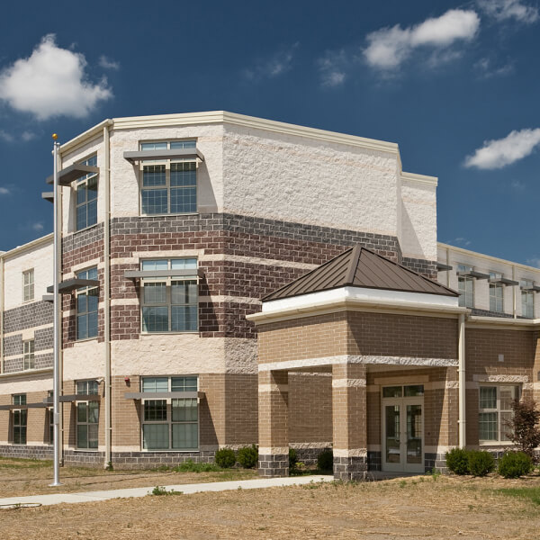 A modern, two-story brick and stone school building with large windows, a small entrance canopy, and a flagpole in front, set against a blue sky with a few clouds.