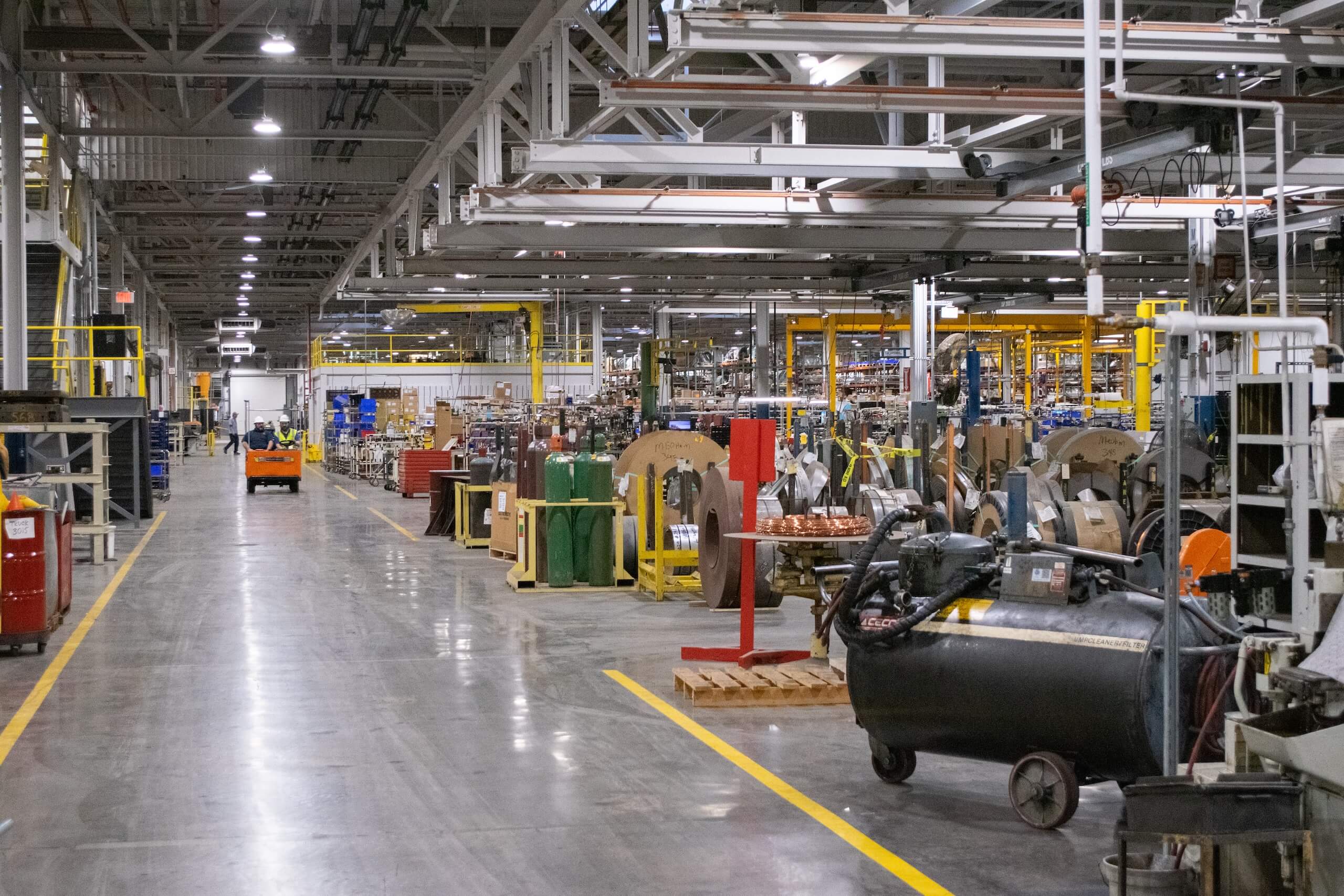Interior of a large industrial factory with machines, equipment, and workstations. A few workers and an orange vehicle are visible in the background. The floor is clean and marked with yellow safety lines.