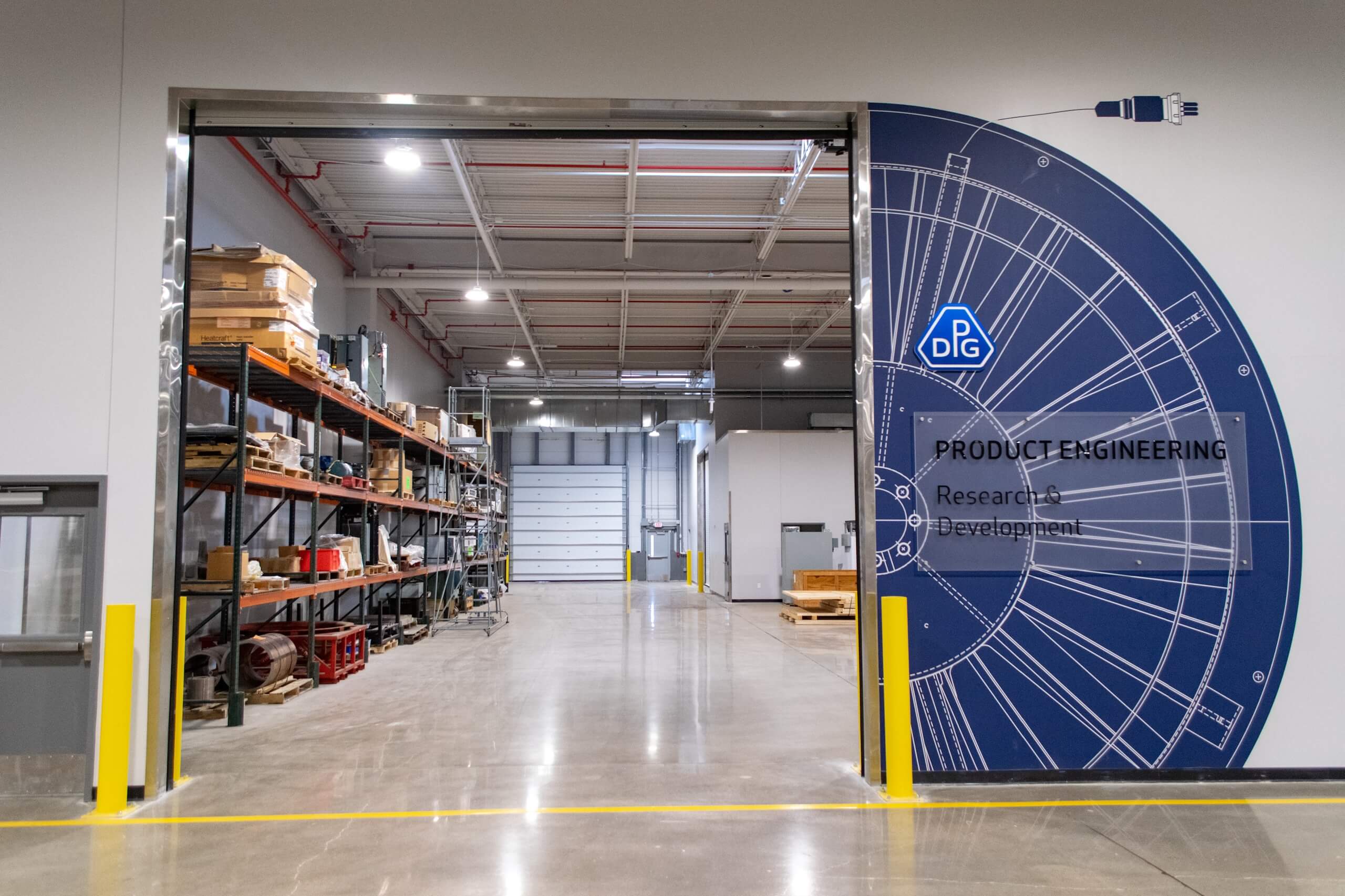 Open industrial workspace with shelves holding boxes and materials, a large blue Product Engineering wall graphic, polished concrete floor, and a partially opened garage door at the back.