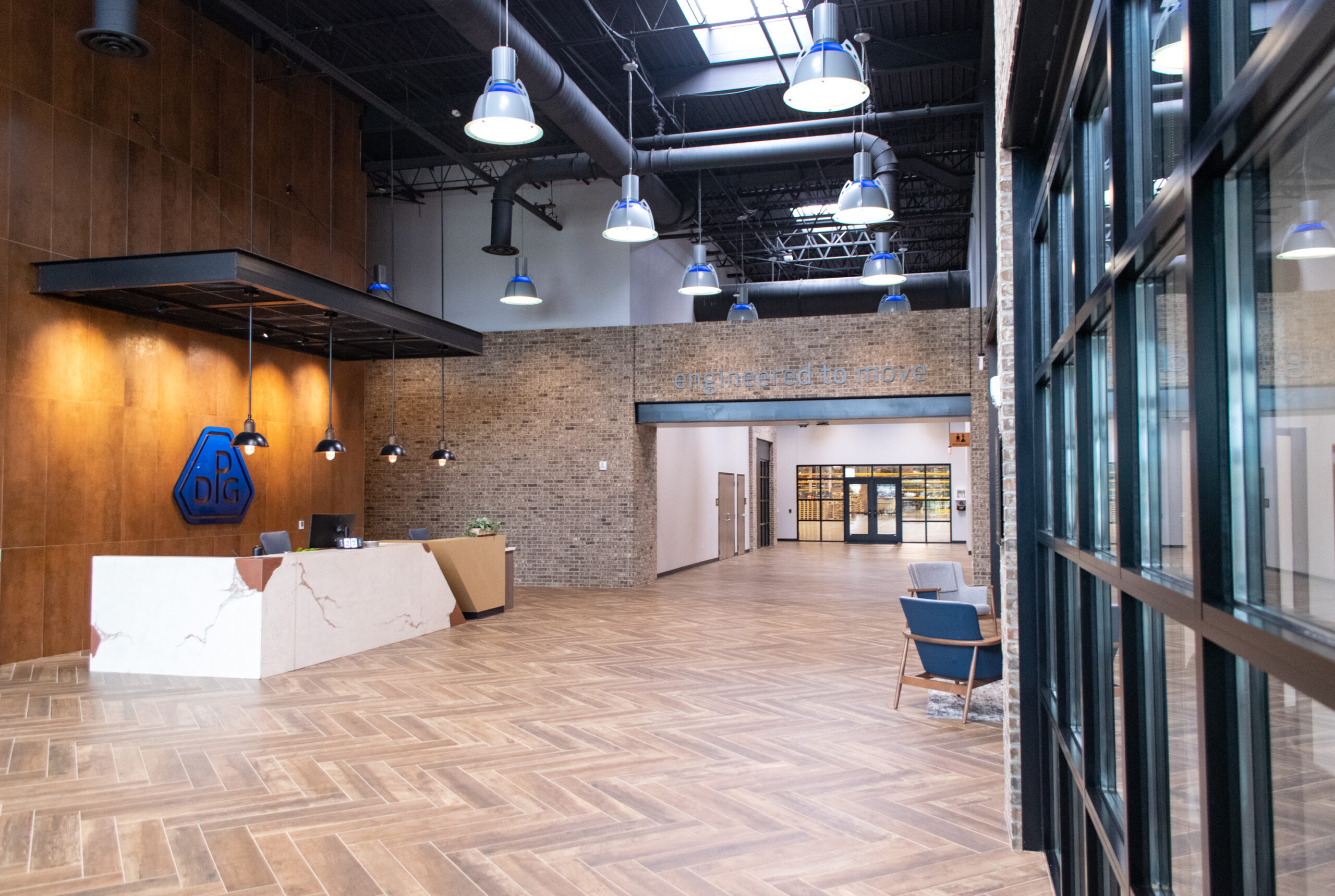 A modern lobby with a white marble reception desk, wood-patterned floors, exposed brick walls, pendant lights, blue accent chairs, and large windows. Double doors lead to another area in the background.