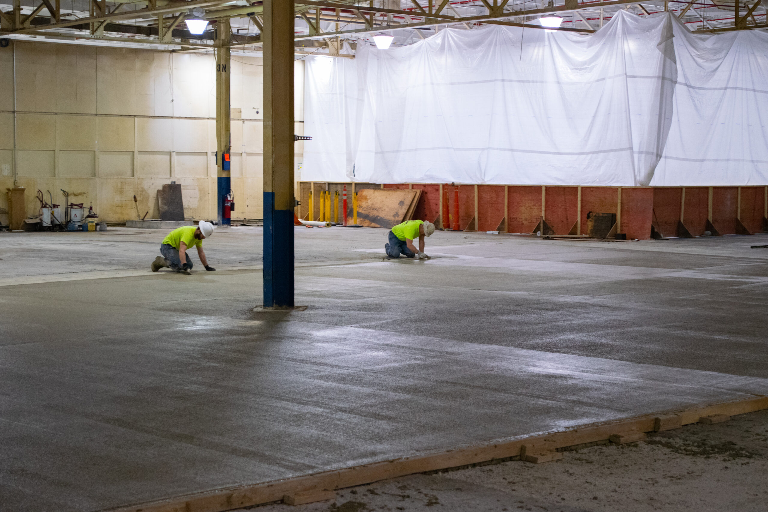 Two construction workers in safety gear kneel on a large concrete floor, smoothing the surface inside a spacious industrial building under construction with a plastic sheet draped in the background.