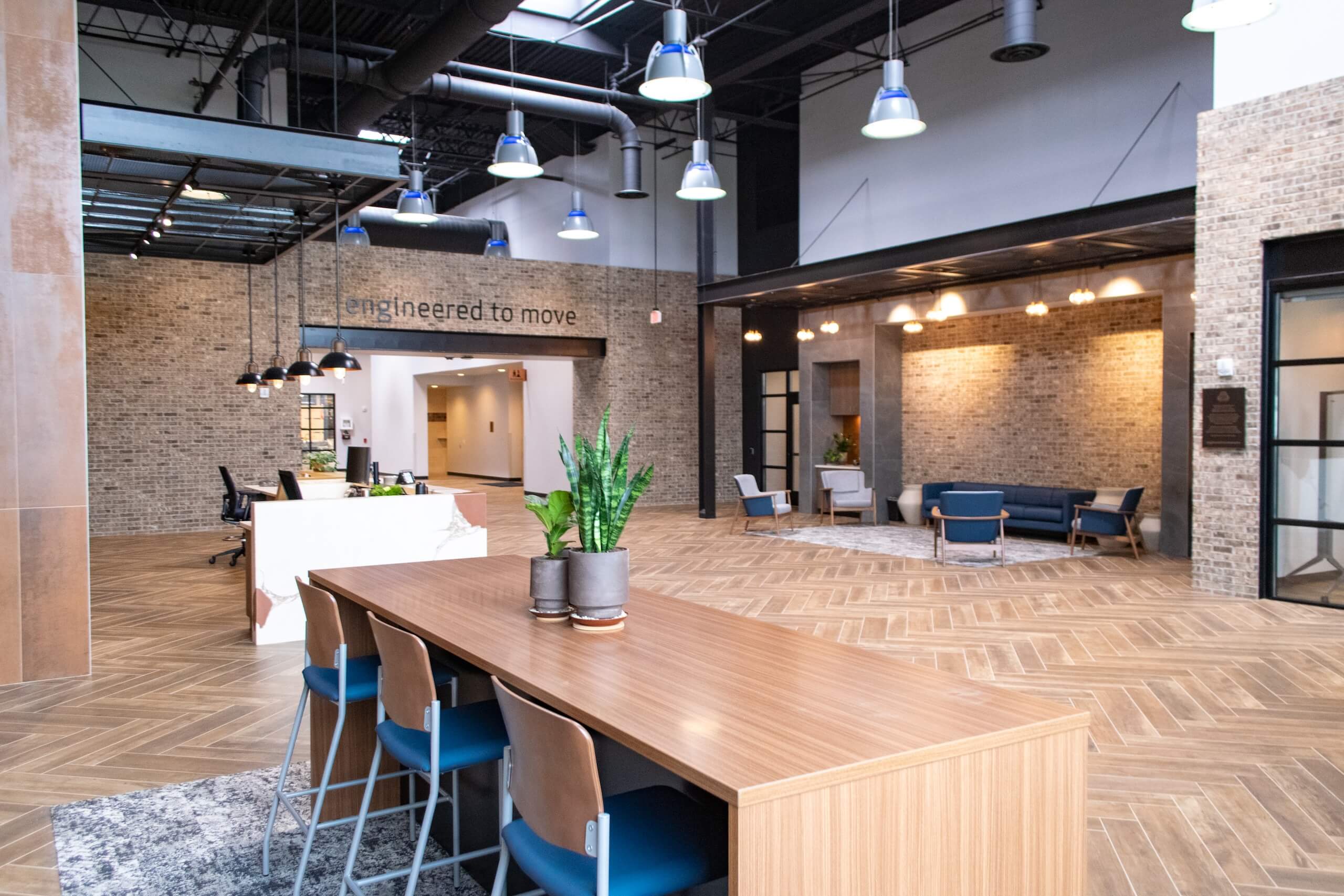 Modern office lobby with wooden floors, indoor plants, seating areas, exposed brick walls, and high ceilings with hanging lights. A sign on the wall reads engineered to move..