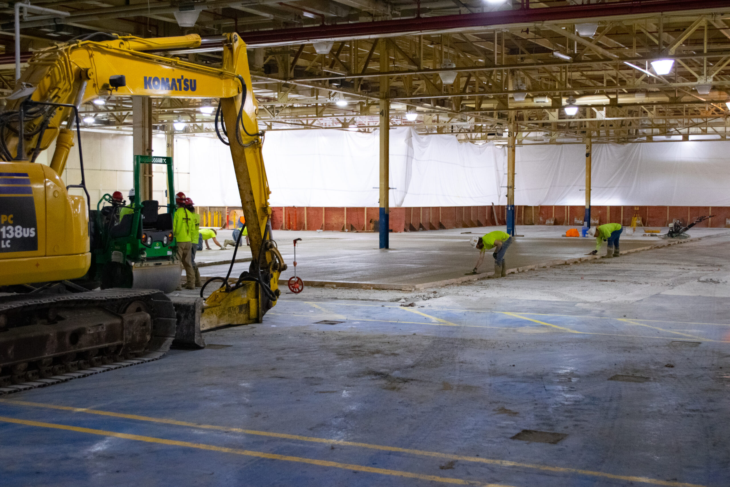 Workers in safety gear use heavy machinery and tools to break up concrete flooring inside a large, partially demolished industrial warehouse.