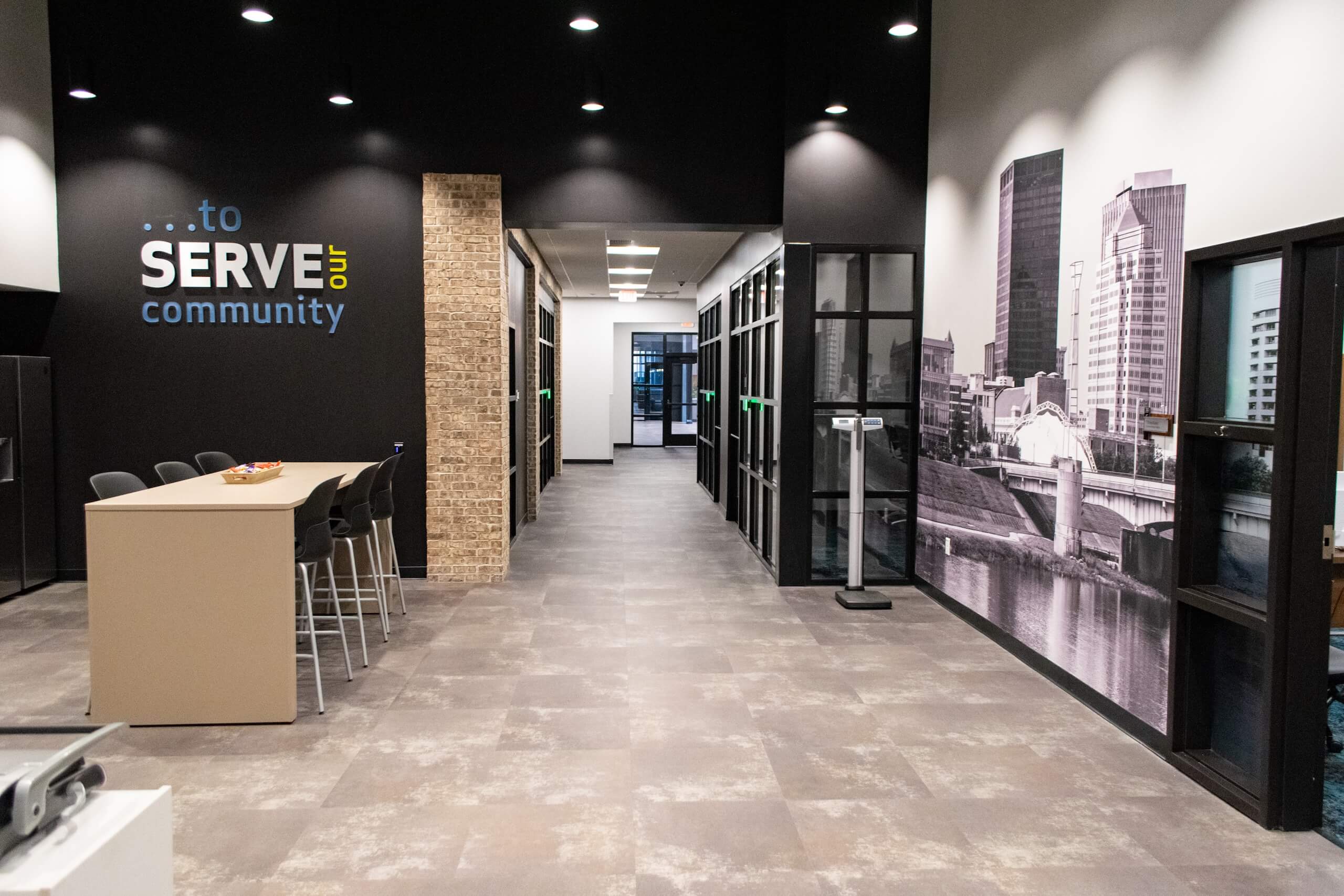 Modern office hallway with glass-walled rooms, a meeting table with chairs, and a large wall mural of a cityscape. A wall displays the words ...to SERVE our community in blue and yellow letters.