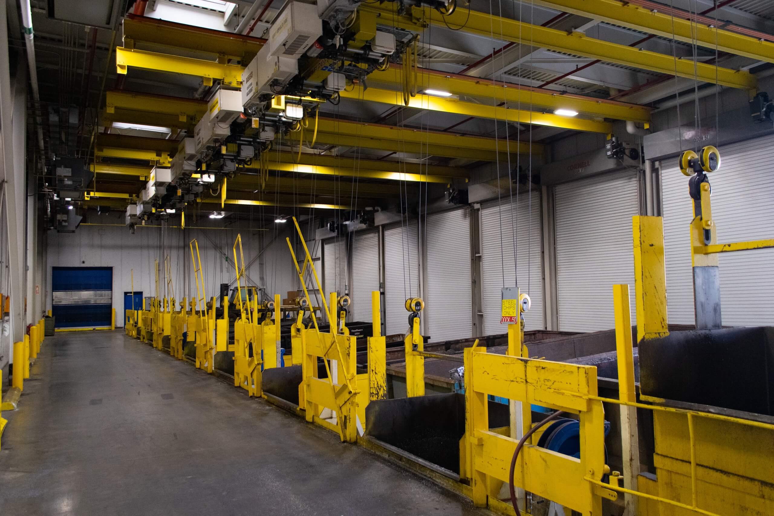 A large industrial facility with yellow overhead cranes, machinery, and safety barriers, featuring concrete floors and large garage doors along one wall. The space appears organized and well-lit with fluorescent lights.