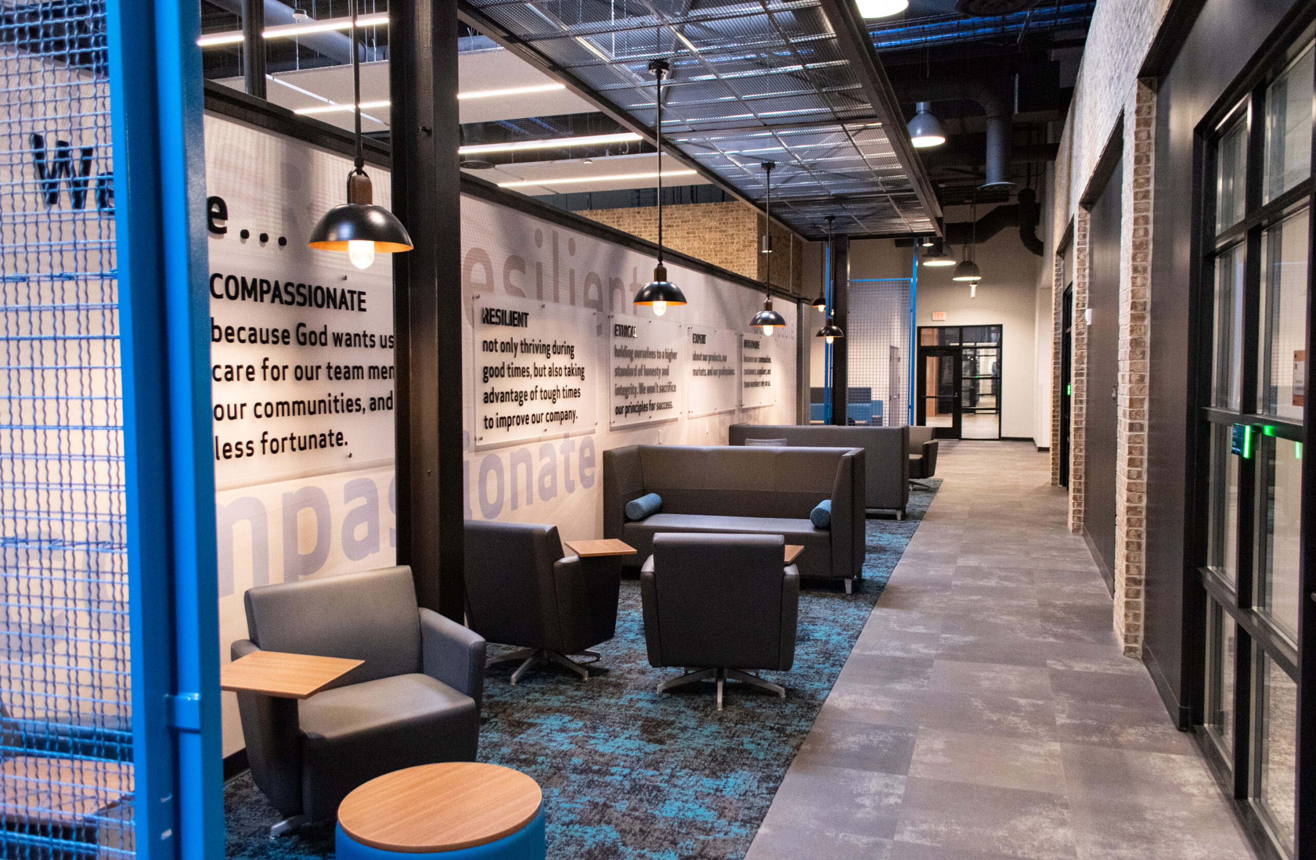 Modern indoor lounge with gray chairs, small tables, and wall art featuring inspirational words. Overhead lights hang from an exposed industrial ceiling. Blue and gray tones dominate the space, which has brick and glass accents.