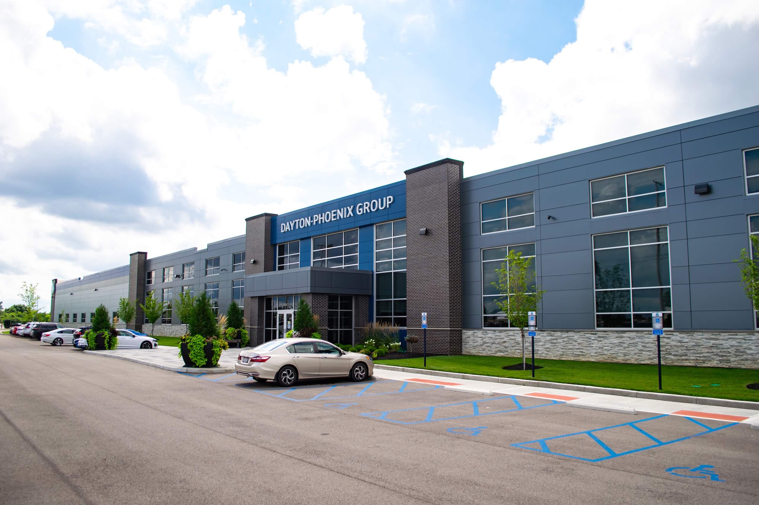 A modern gray office building with large windows, labeled Dayton-Phoenix Group. Several cars are parked outside, and handicap parking spaces are visible in the foreground. Trees and a cloudy sky are in the background.