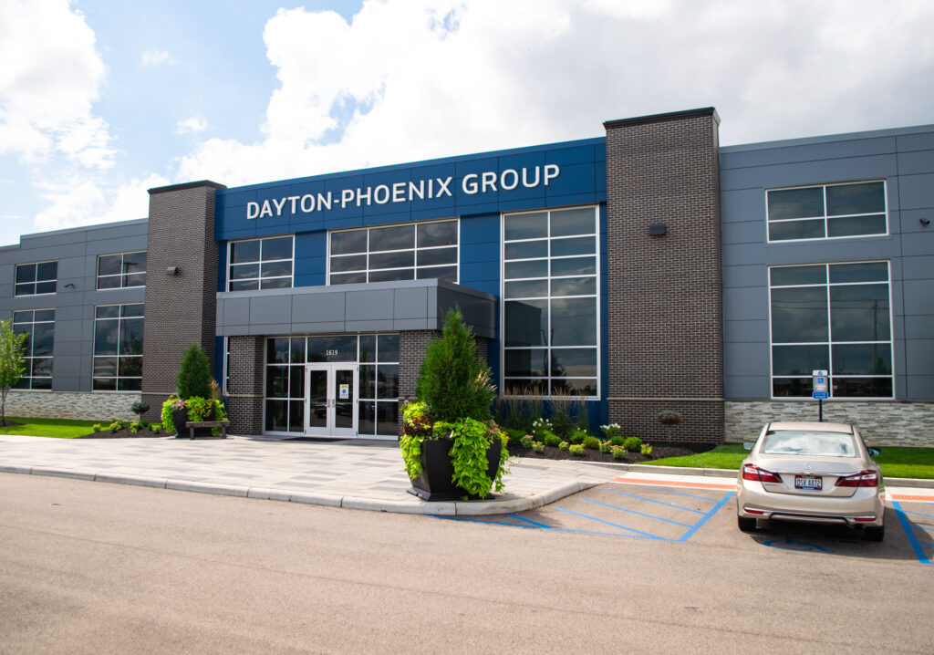 Modern office building with a sign reading Dayton-Phoenix Group, large windows, landscaped greenery, and a car parked in an accessible parking space in front.