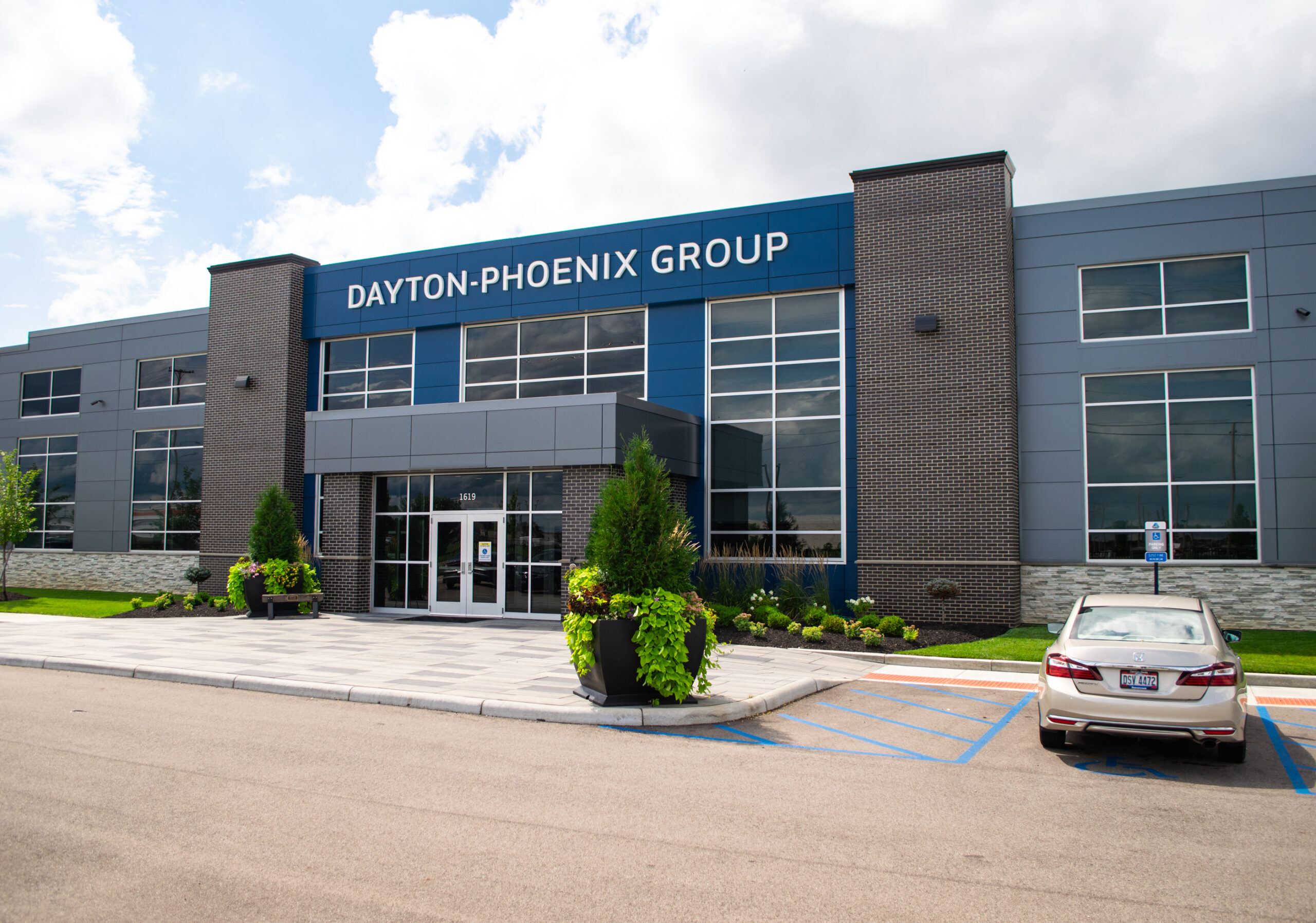 Modern office building with a sign reading Dayton-Phoenix Group, large windows, landscaped greenery, and a car parked in an accessible parking space in front.