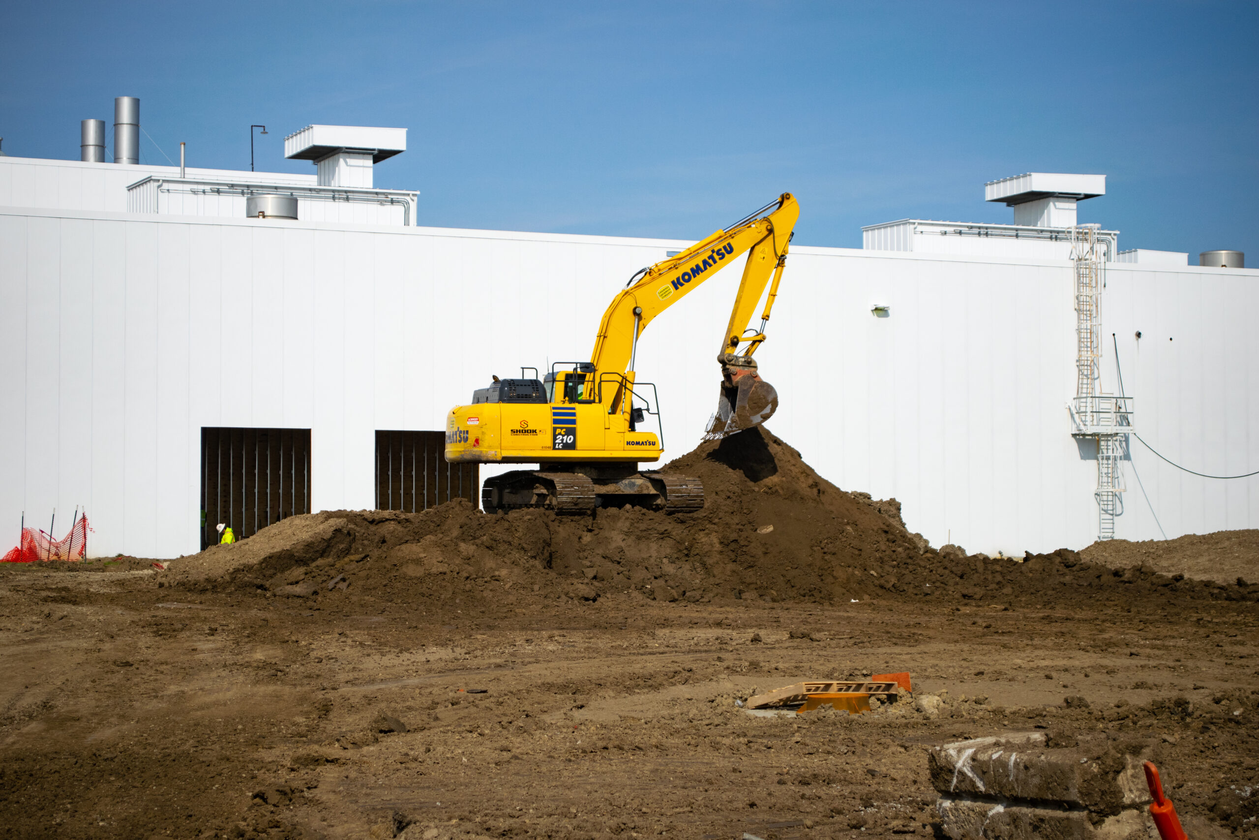A yellow excavator moves a pile of dirt at a construction site in front of a large, white industrial building under a clear blue sky.