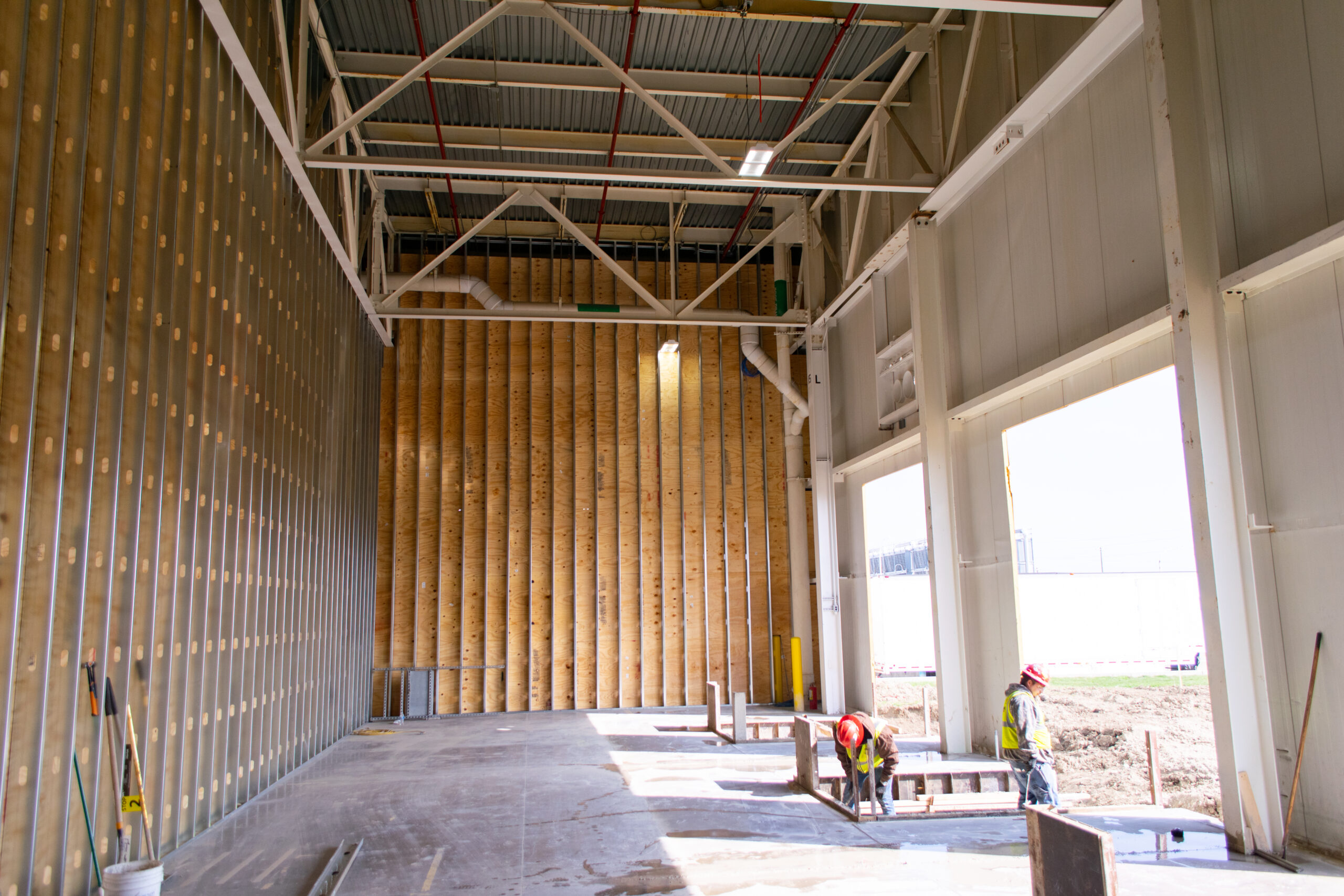 Partially constructed industrial building interior with exposed framing, tall wooden wall, and sunlight streaming in. Two construction workers in safety gear stand near tools and materials on the right side.