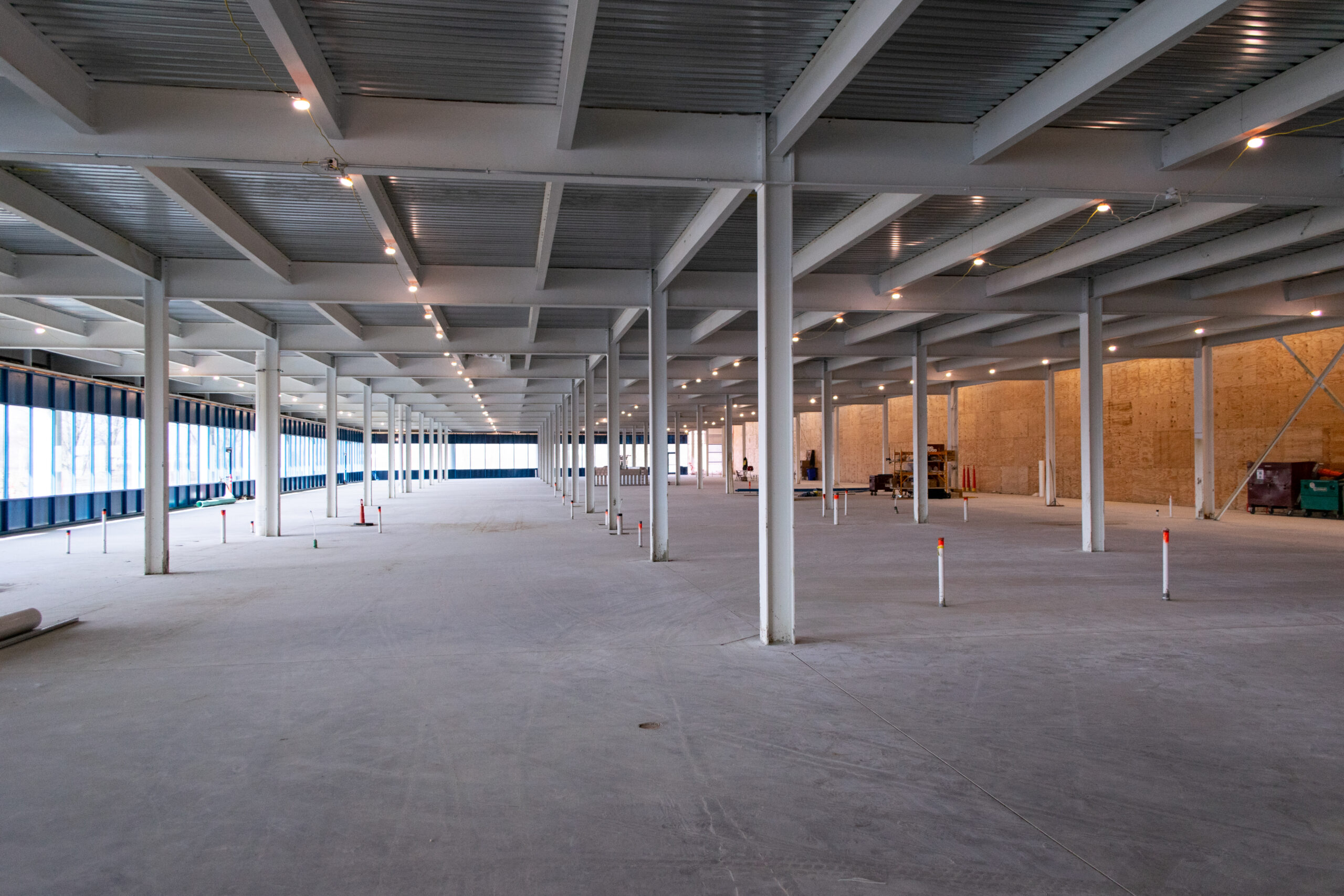 Large, empty warehouse under construction with exposed beams, unfinished flooring, and some scattered construction materials and equipment. Ceiling lights are on, and pipes extend from the ground. Natural light enters from the left windows.