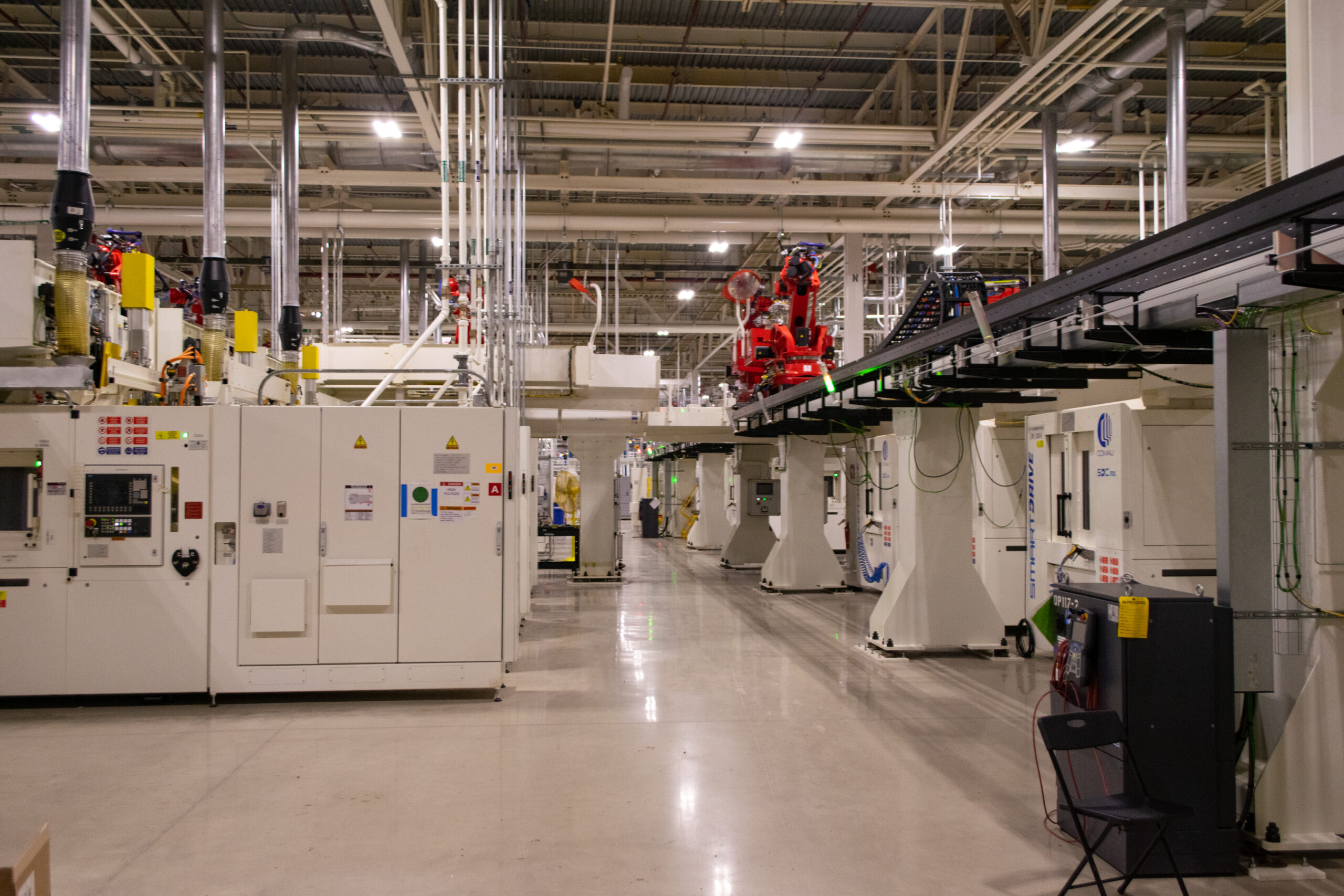 A clean, well-lit industrial factory floor with white machinery, cables, red robotic arms, control panels, and an overhead conveyor system. Empty black chairs are placed along the right side of the image.