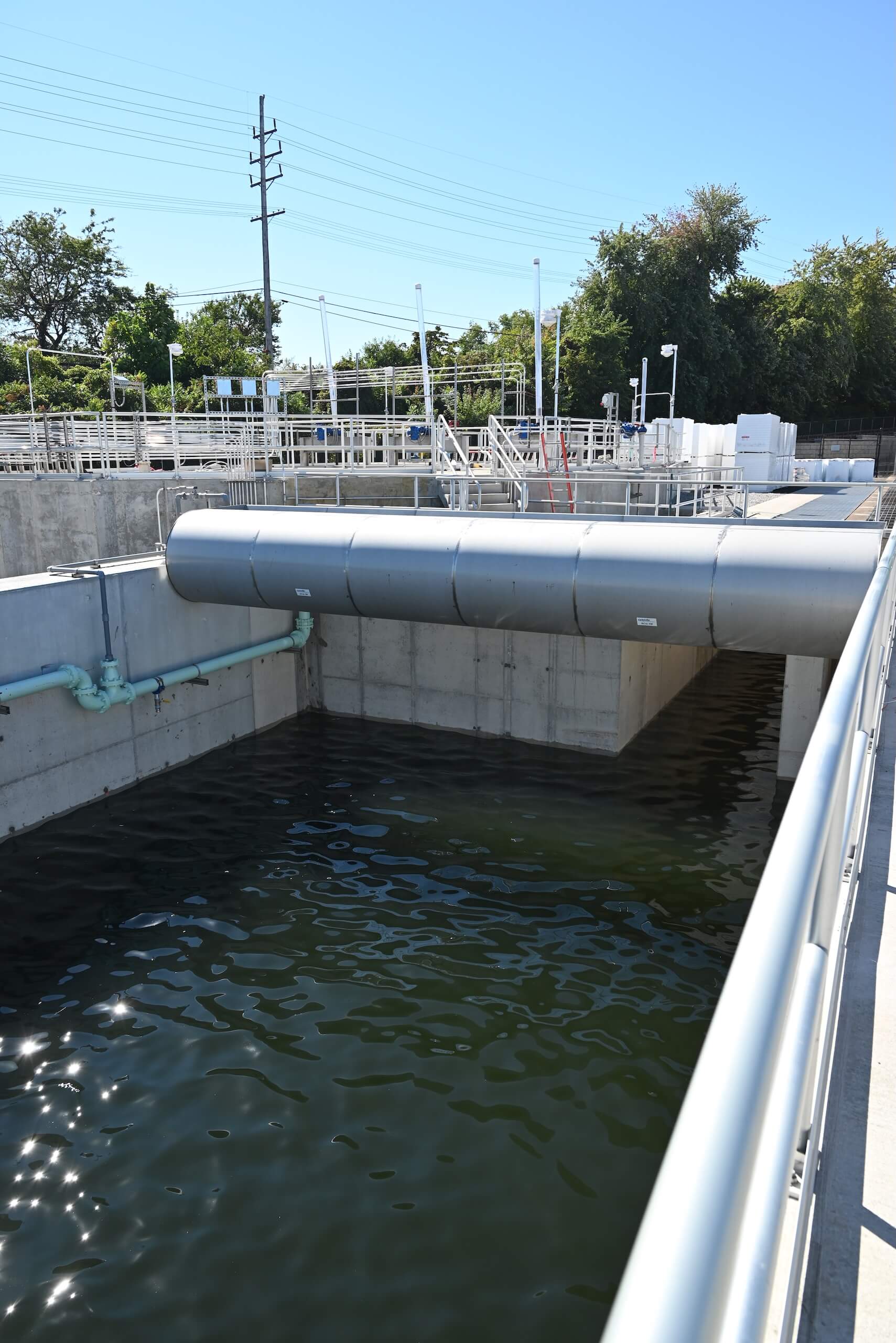 A large outdoor water treatment facility with concrete basins, metal railings, and pipes, filled with dark water under a clear blue sky. Trees and utility poles are visible in the background.