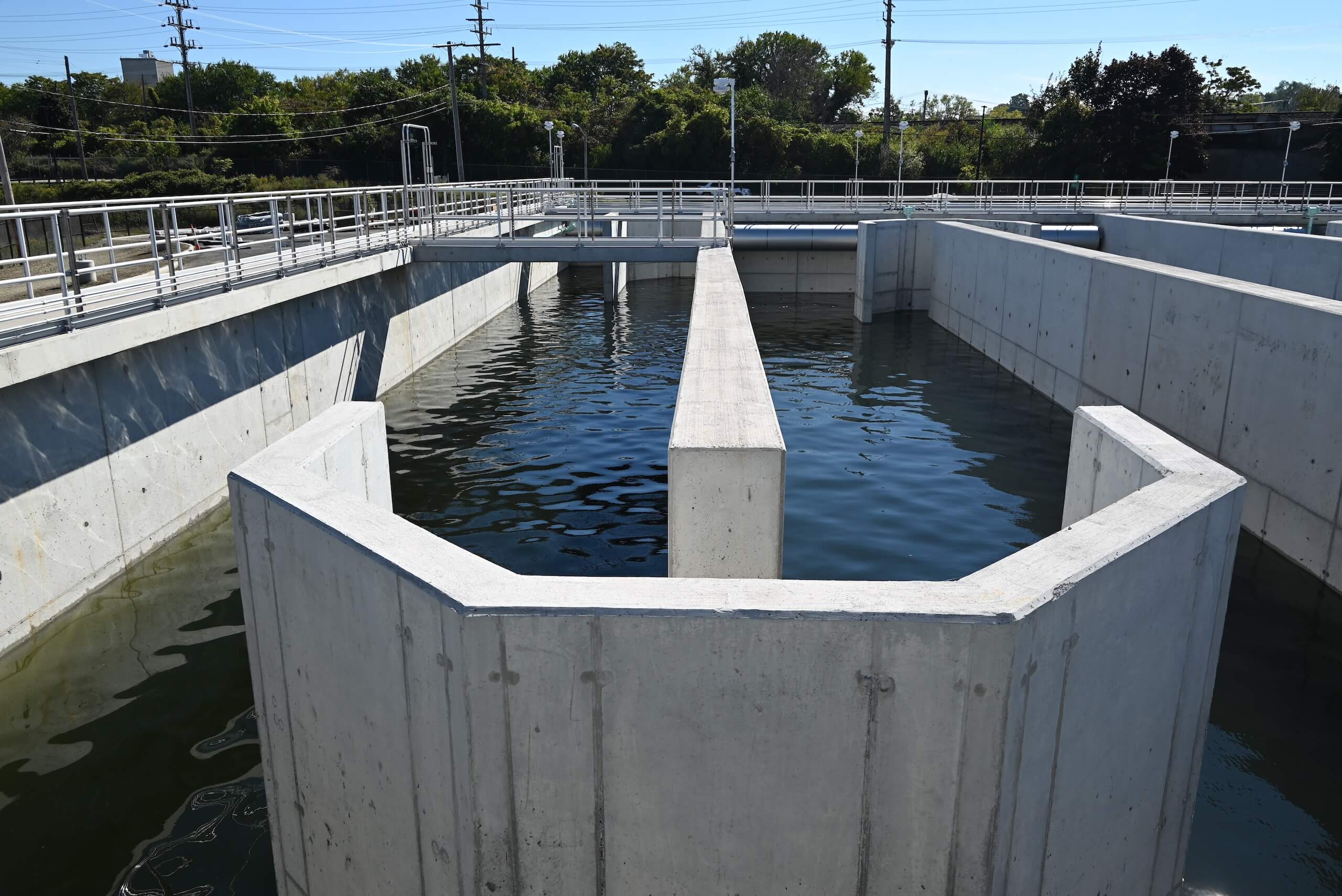 A large concrete water treatment basin filled with water, surrounded by railings and trees in the background under a clear sky.