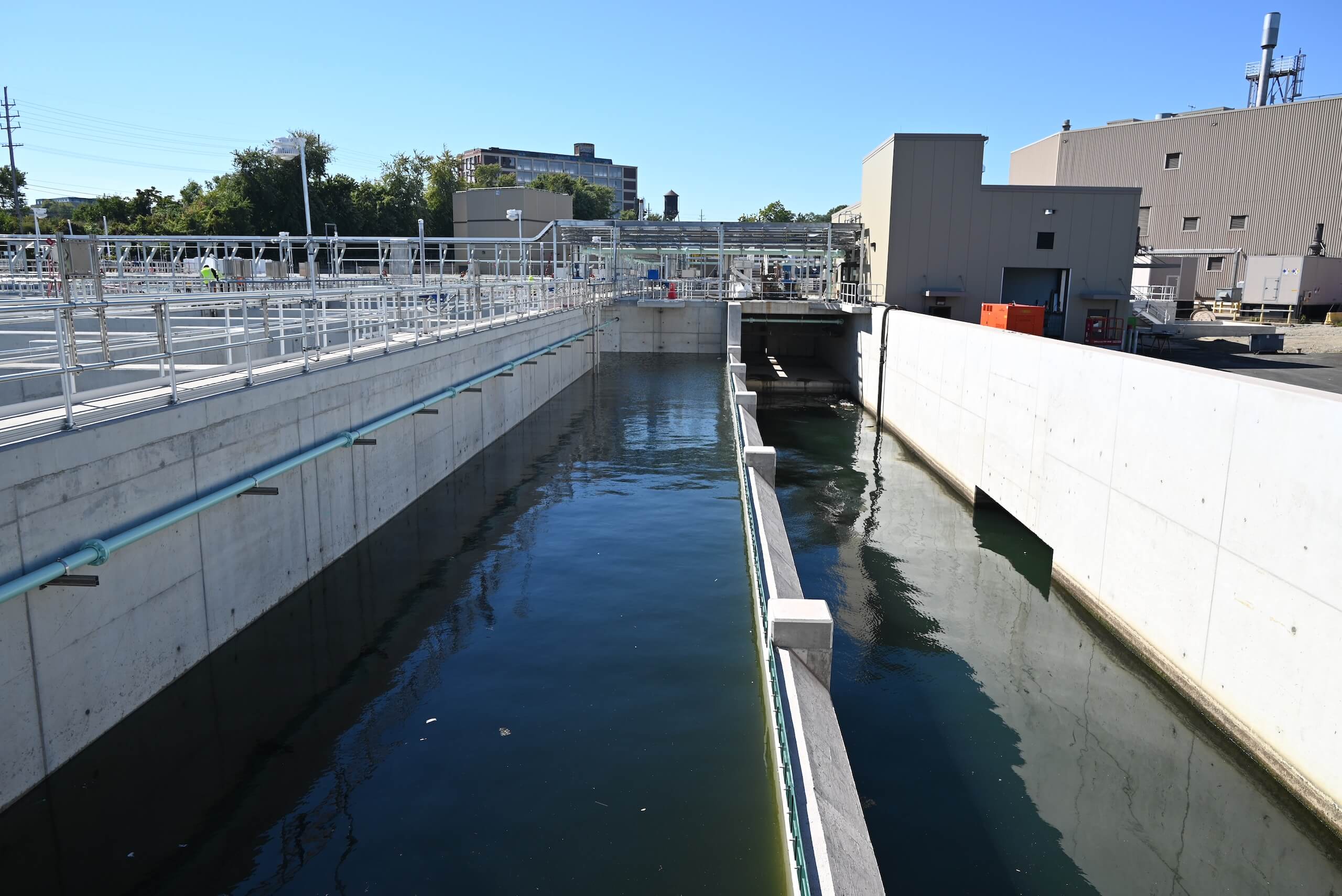 A water treatment facility with concrete channels carrying water, surrounded by industrial buildings and pipes, under a clear blue sky.