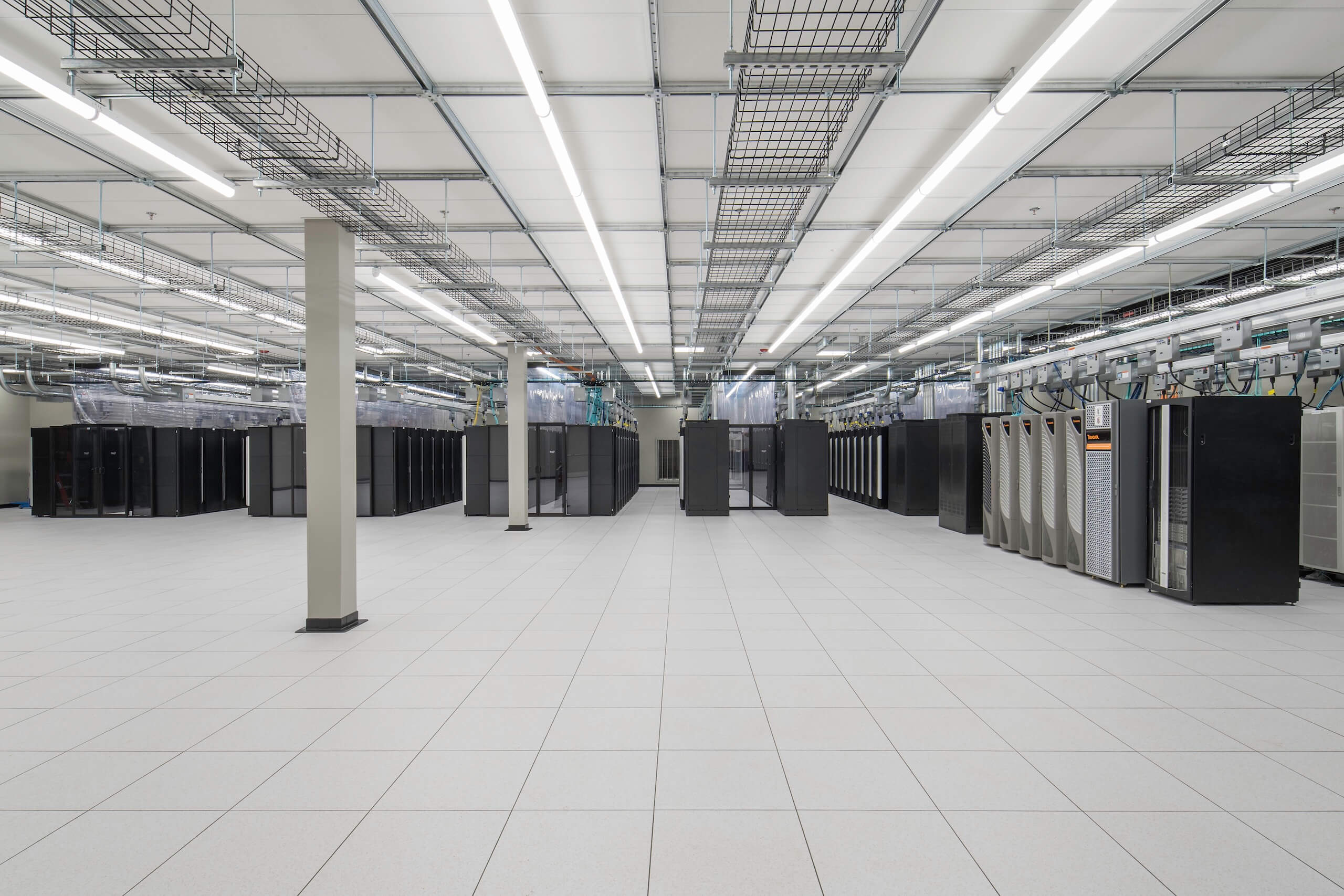 Large, clean data center with rows of black server racks, white tiled floor, overhead cable trays, and bright fluorescent ceiling lights. The room is spacious and modern.