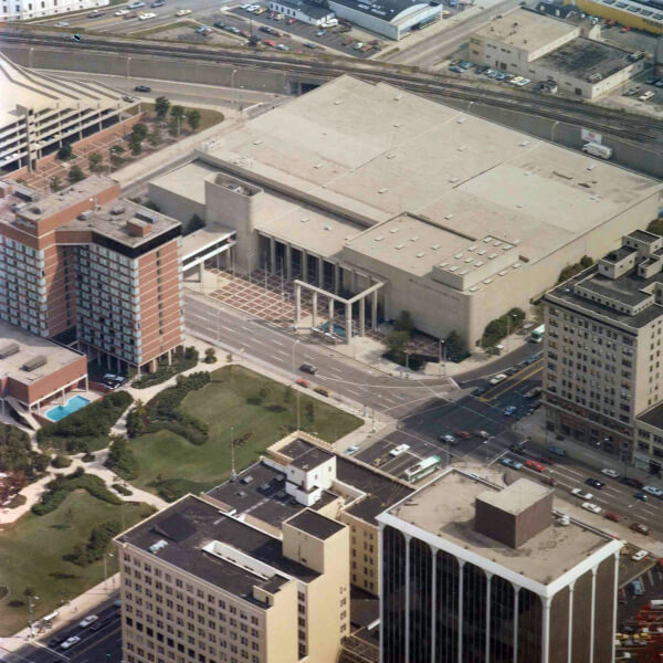 Aerial view of a city block featuring a large, beige convention center, surrounding office buildings, a pool, streets, and a small green park area with sidewalks. Cars are visible along the roads.