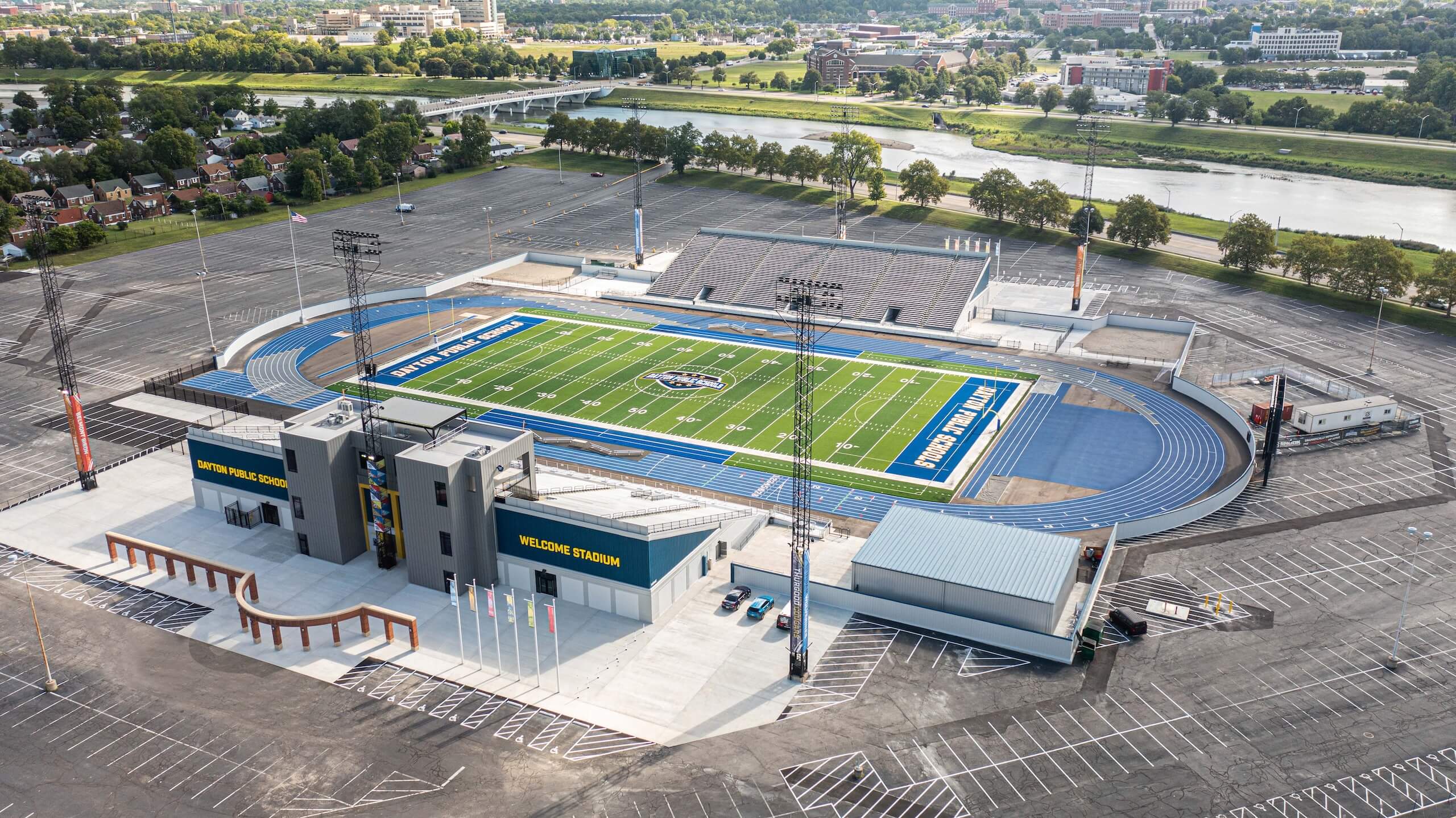 Aerial view of an empty outdoor stadium with a green football field, blue running track, Welcome Stadium signs, seats, parking lot, and a river with trees in the background.
