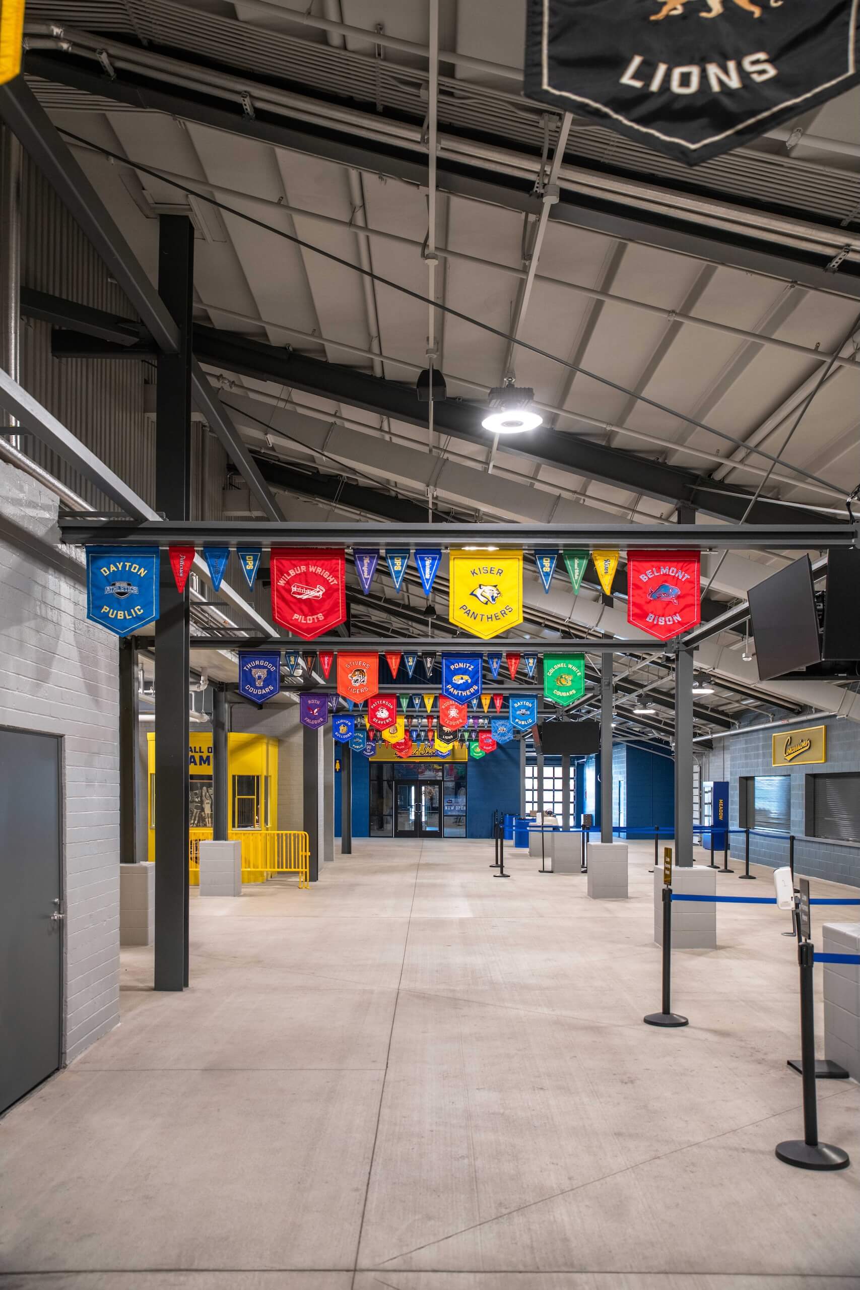 A spacious indoor hallway with colorful triangular and shield-shaped school banners hanging from the ceiling. Concrete floors, yellow railings, and retractable queue barriers are visible. The area appears clean and well-lit.