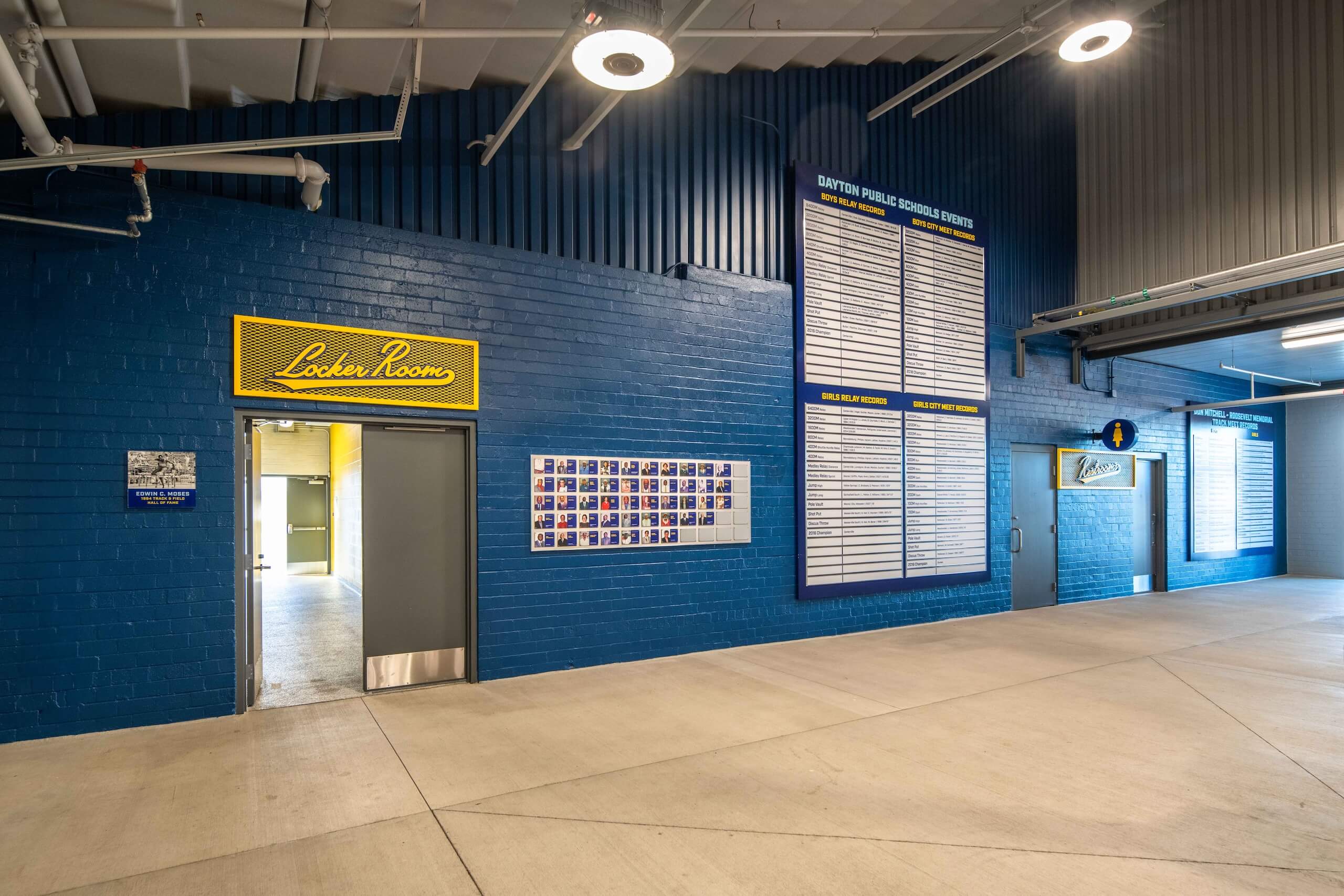 A hallway with blue walls features a Locker Room sign, a wall of photos, a schedule board, and open doors leading to a lit interior. Overhead lights and concrete floors create a clean, modern look.