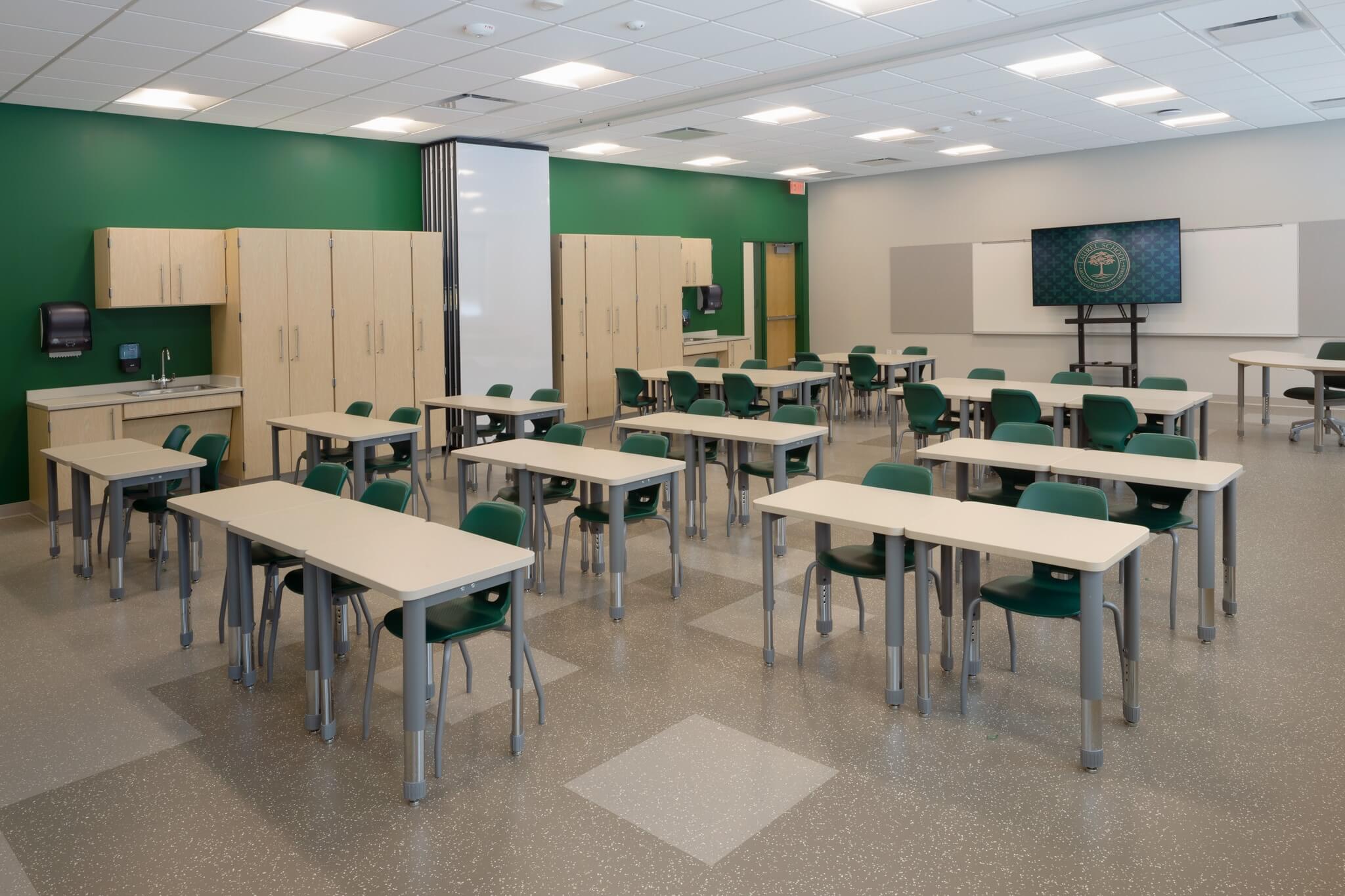 A modern classroom with green chairs, light-colored desks, cabinets, a sink, and a large display screen. The room is spacious, well-lit, and arranged for multiple students.