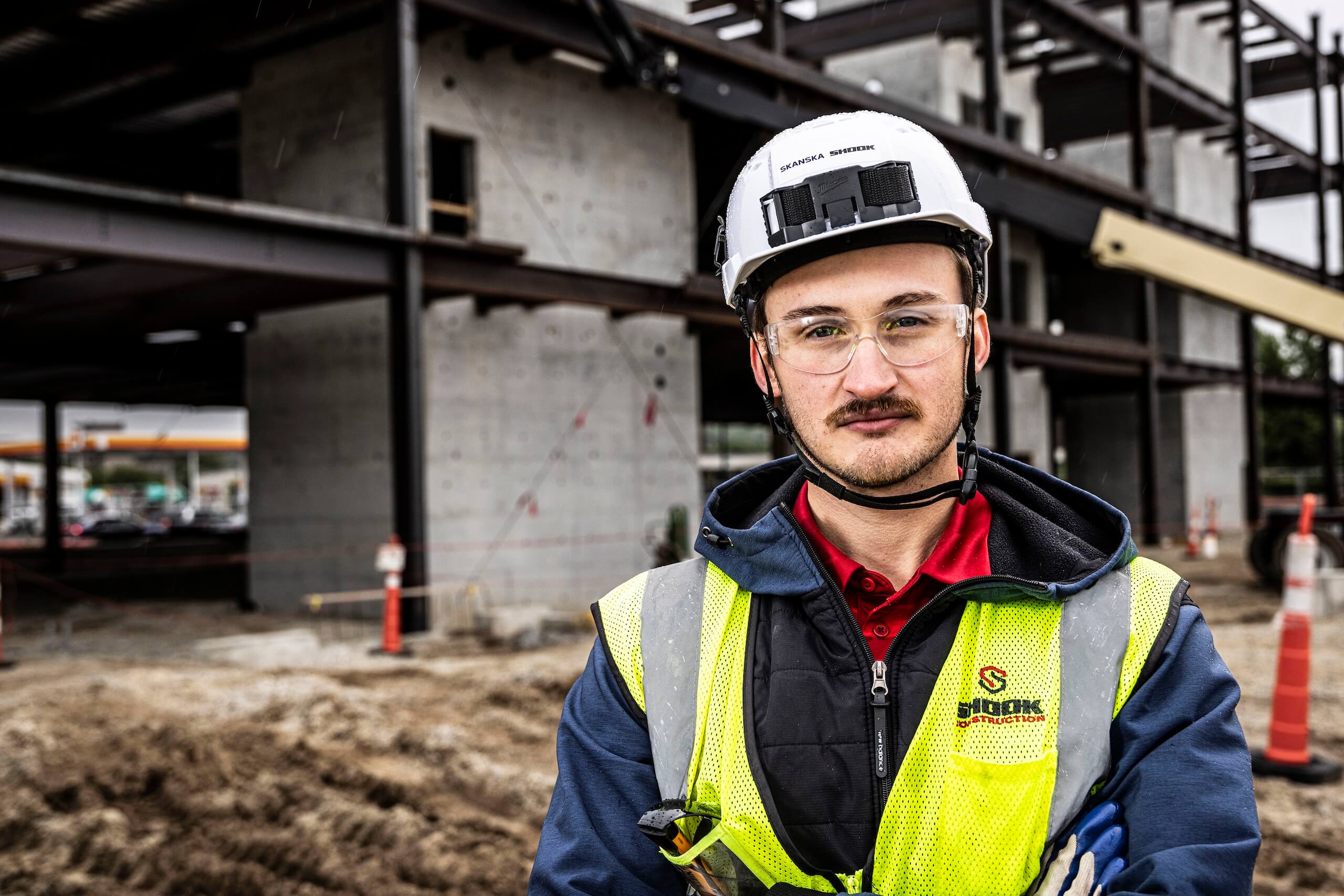 A construction worker wearing a hard hat, safety glasses, and a reflective vest stands confidently at a building site with unfinished concrete and steel structures in the background.