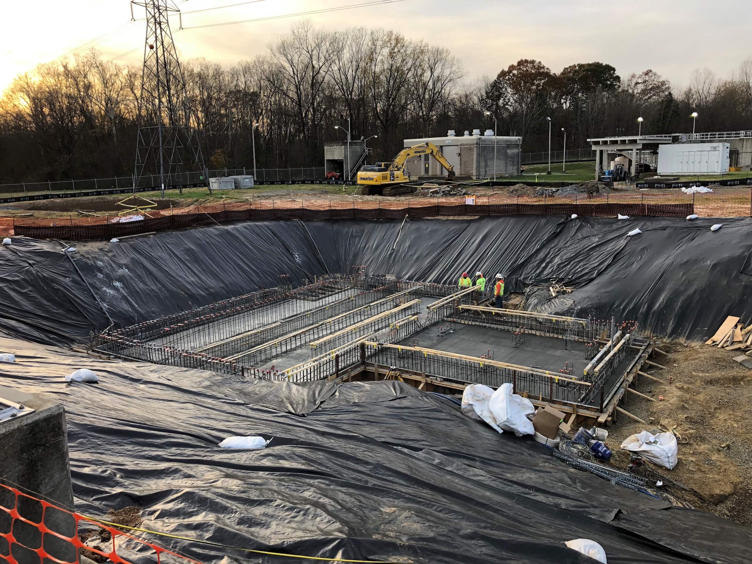 A construction site with a large rectangular pit lined with black plastic, rebar structures, and several workers in safety vests. Machinery and trees are visible in the background.